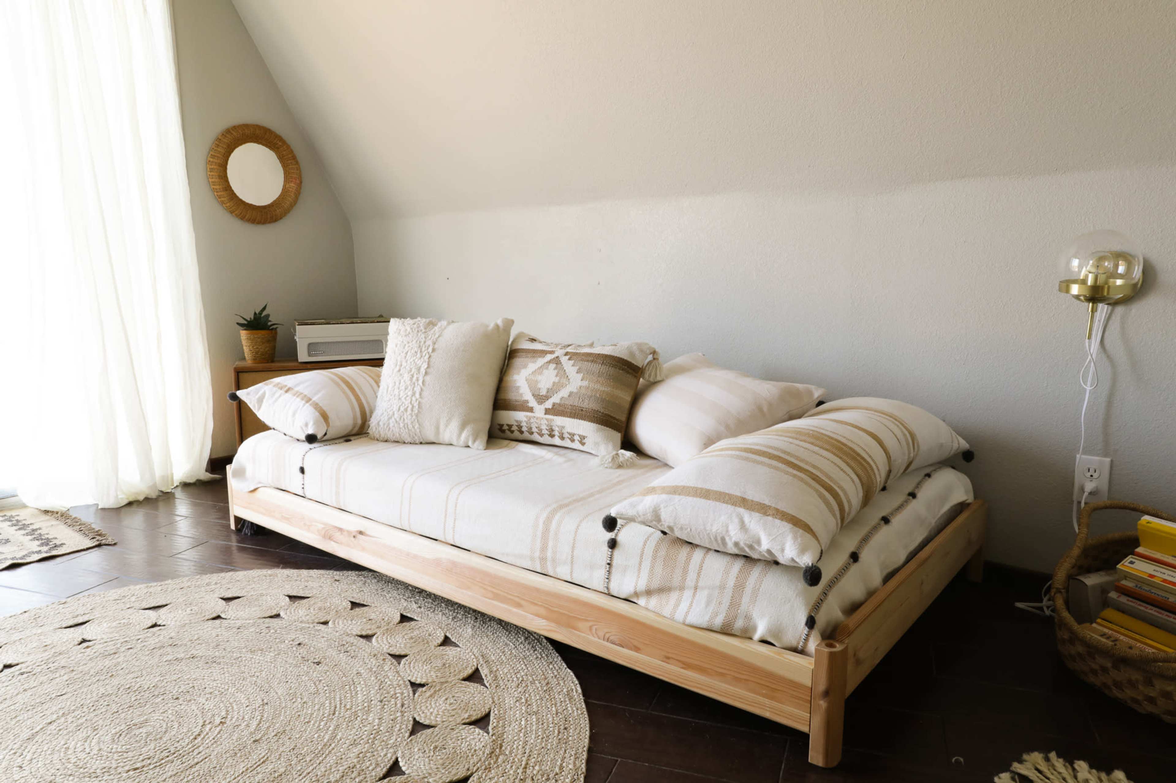 A cozy seating area with a low wooden bed frame adorned with various patterned pillows, next to a round woven rug and a window covered by sheer curtains.