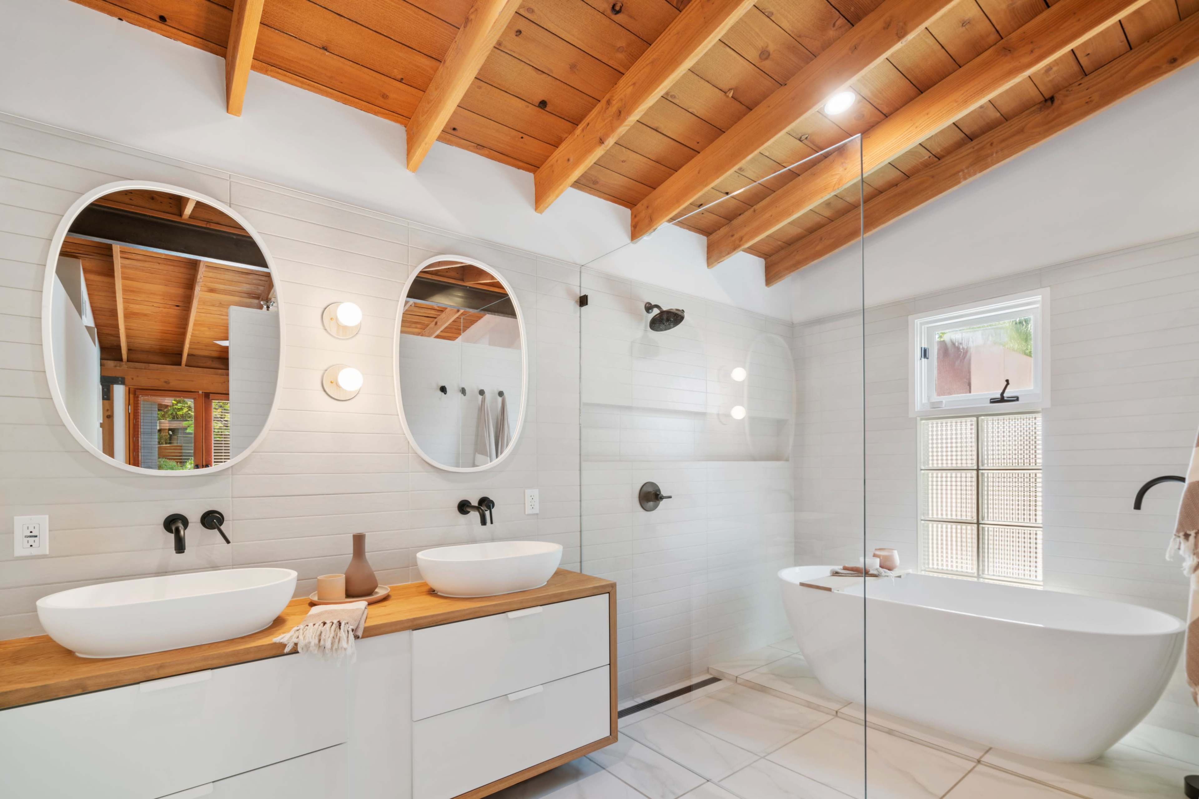 The image shows a modern bathroom featuring dual round mirrors above a wooden countertop with two sinks, a freestanding bathtub, and a glass shower enclosure.