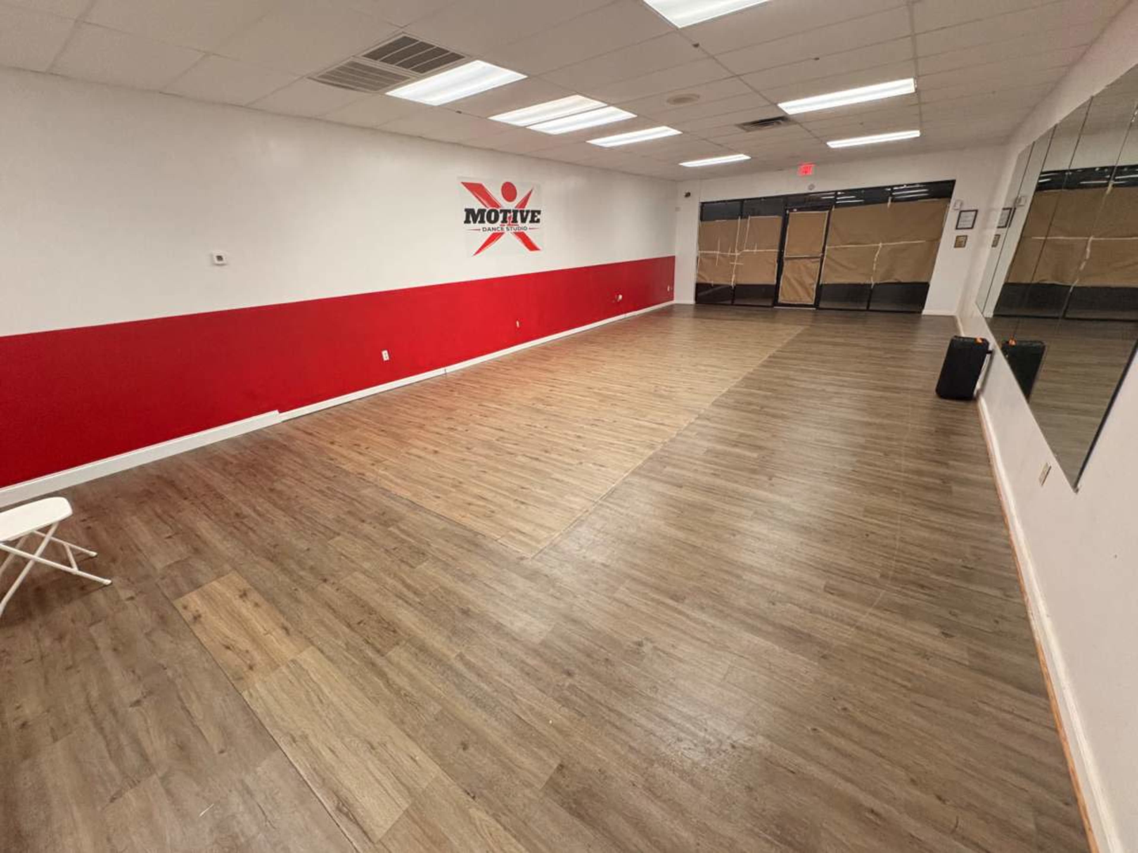 The image shows an empty dance studio with a red and white wall, wooden flooring, and large mirrors along one side.