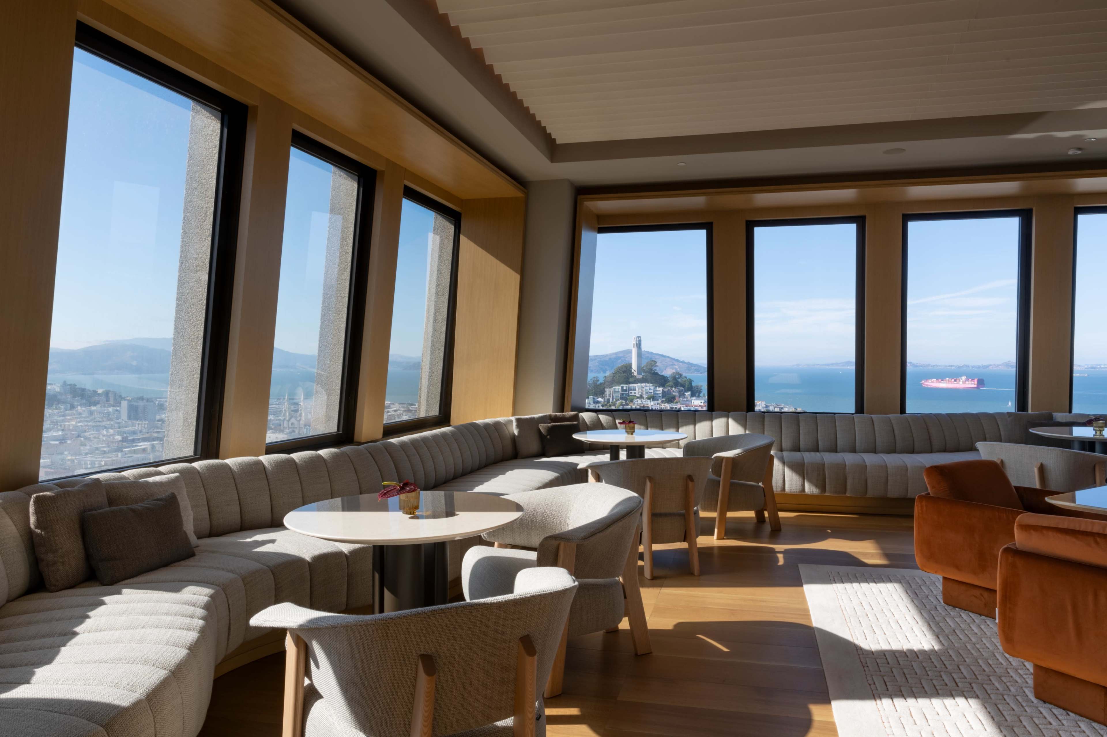 The image shows a modern lounge with large windows offering a view of the water and a distant landmark, featuring comfortable seating arrangements and wooden accents.
