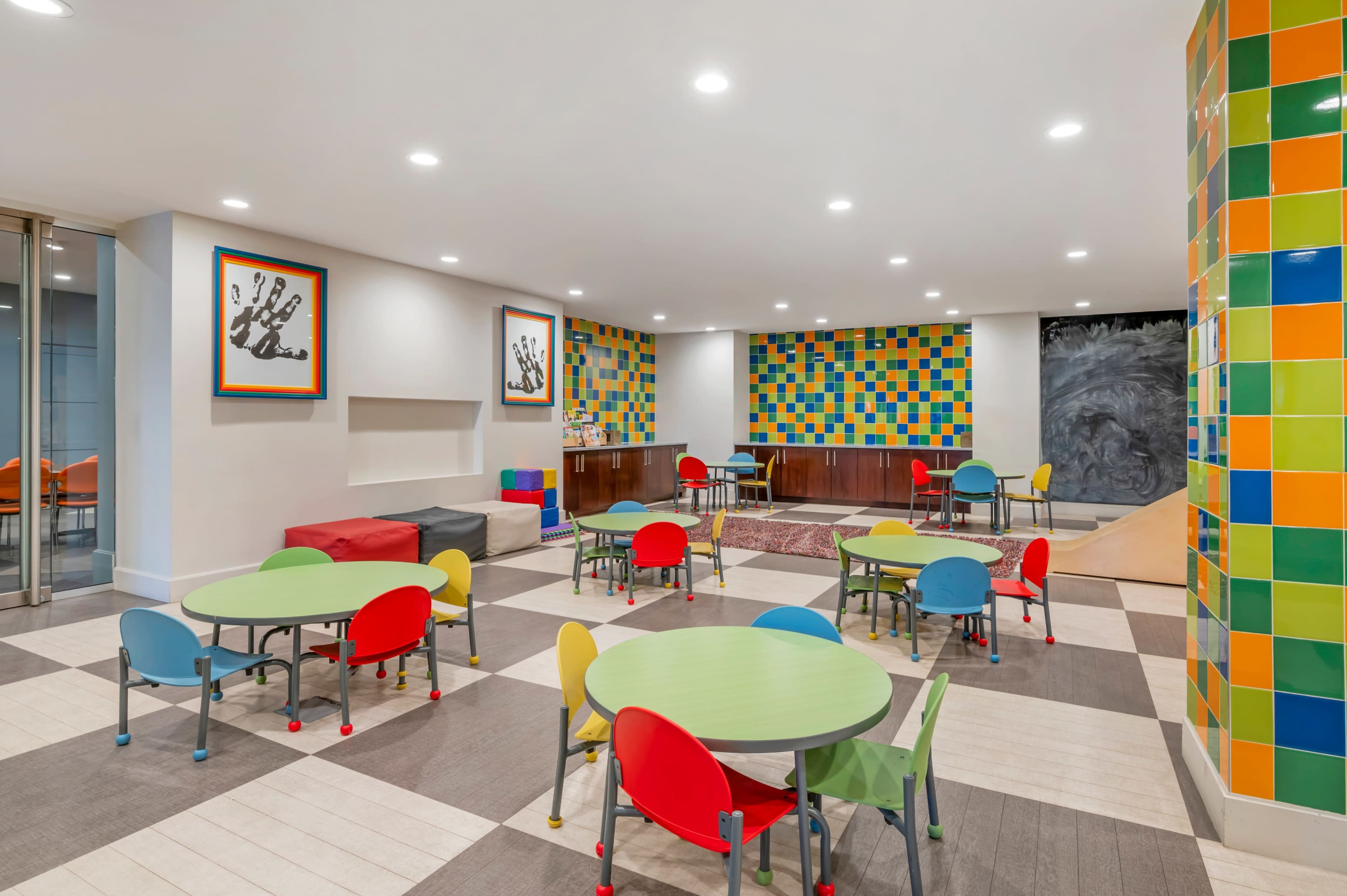 The image shows a brightly colored playroom with various tables and chairs arranged, featuring a colorful tiled wall and artwork.