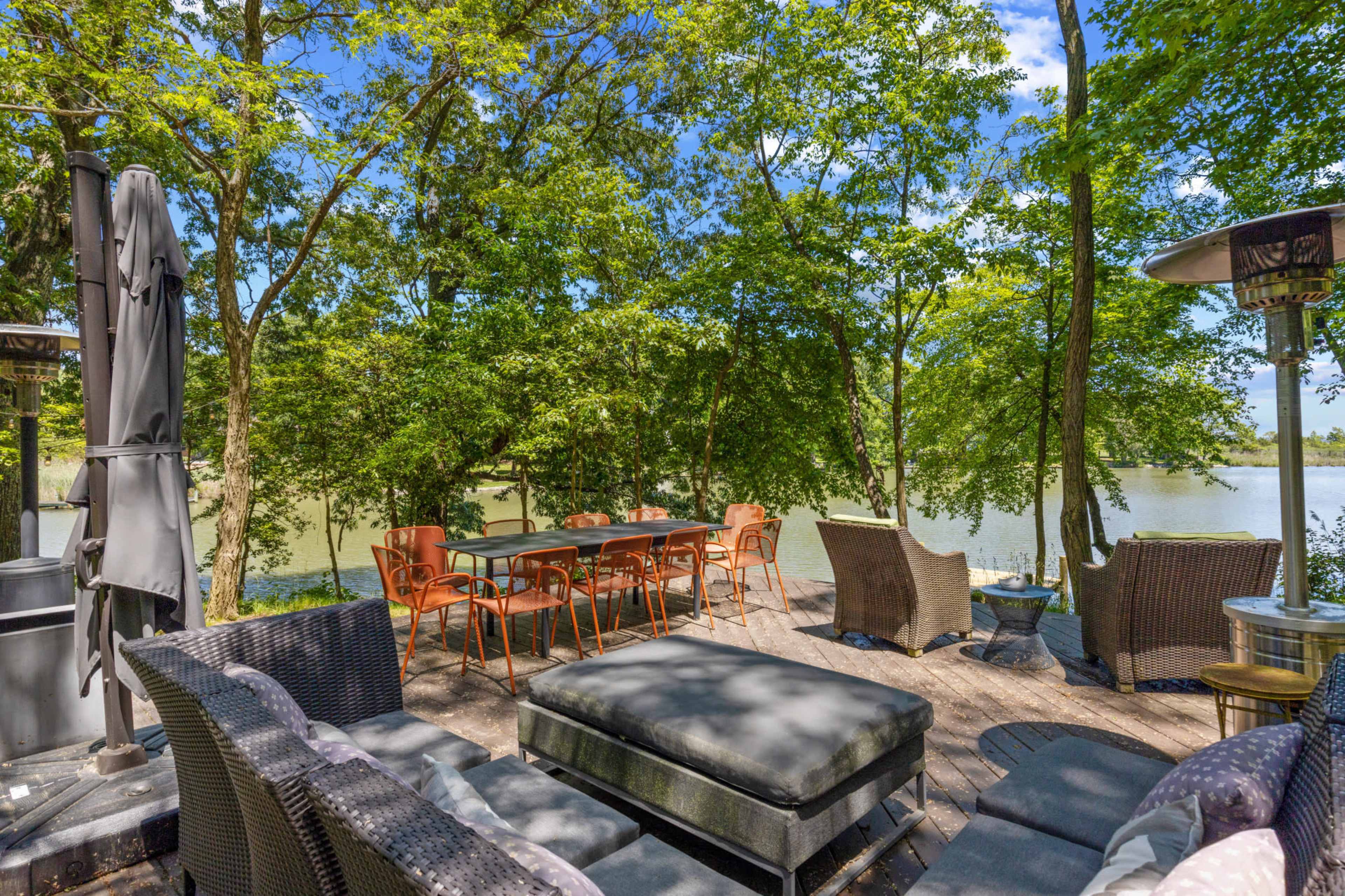 The image shows a wooden deck by a body of water, furnished with various seating arrangements and surrounded by lush greenery.