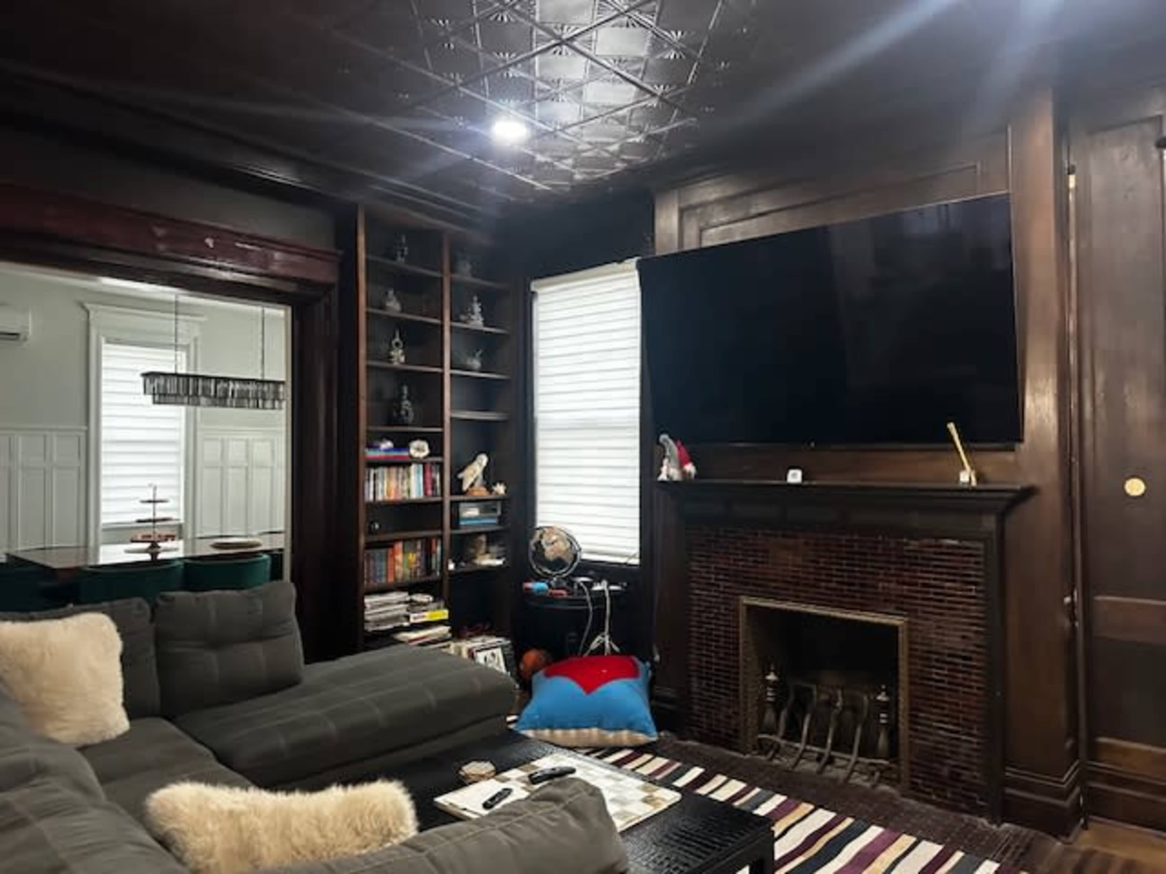 The image shows a living room featuring a dark wood-paneled wall with a large flat-screen TV above a fireplace, a gray sectional sofa with decorative pillows, and bookshelves filled with books and decorative items.
