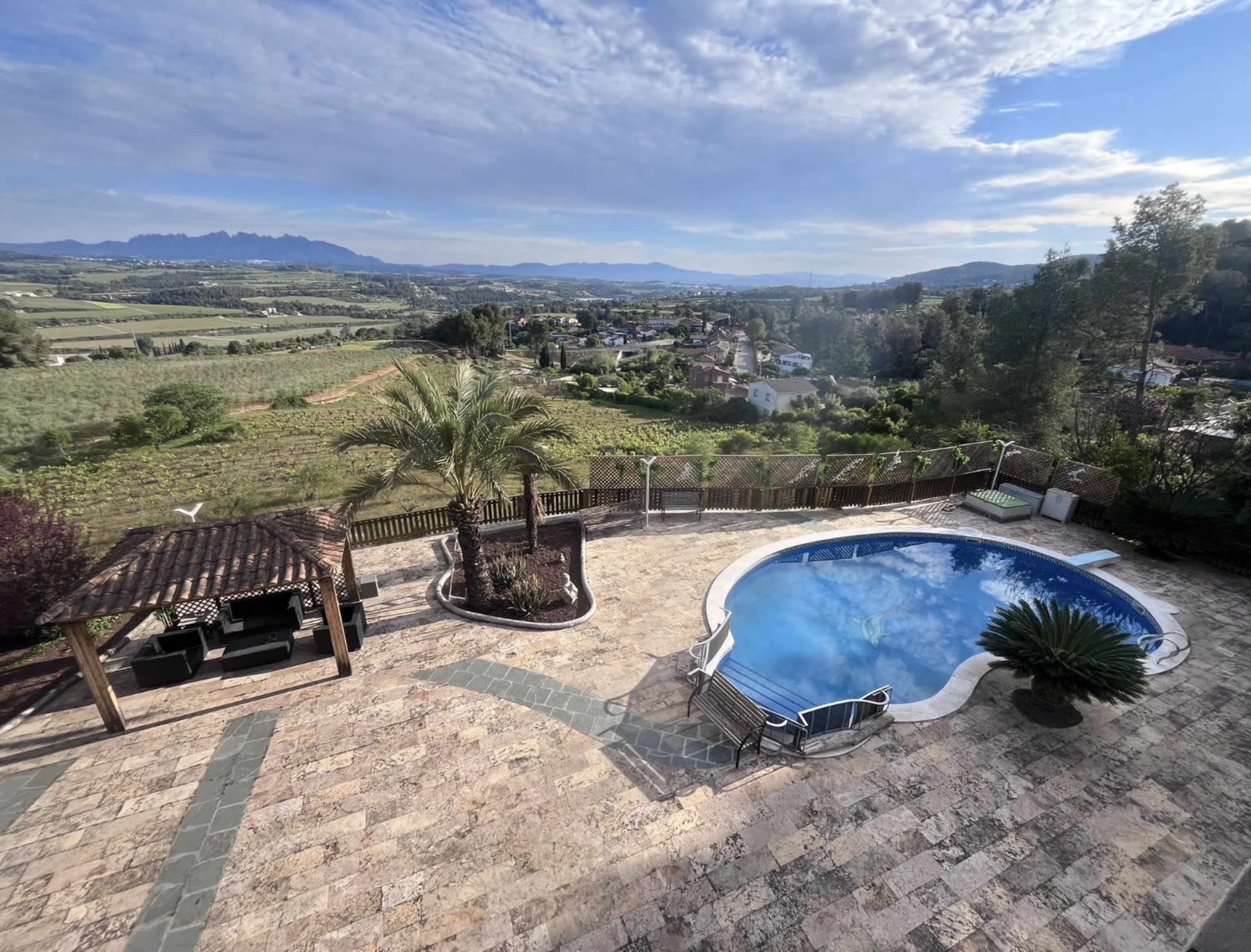 The image shows a scenic view of a property featuring a swimming pool, palm trees, and a patio area overlooking a green landscape and distant mountains.