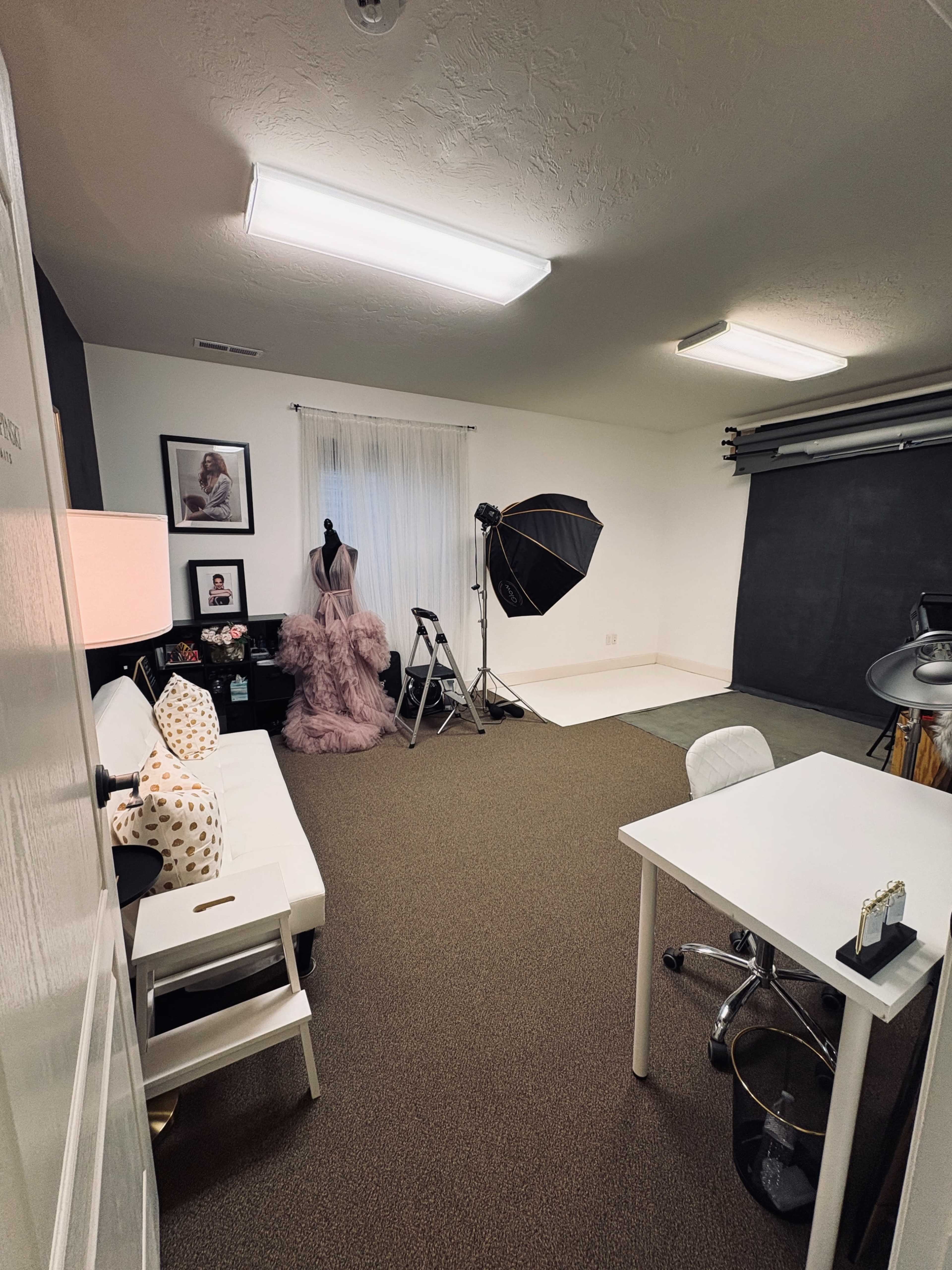 The image shows a well-lit photography studio featuring a white desk, a sofa with decorative cushions, a mannequin dressed in a garment, and a backdrop setup with softboxes.