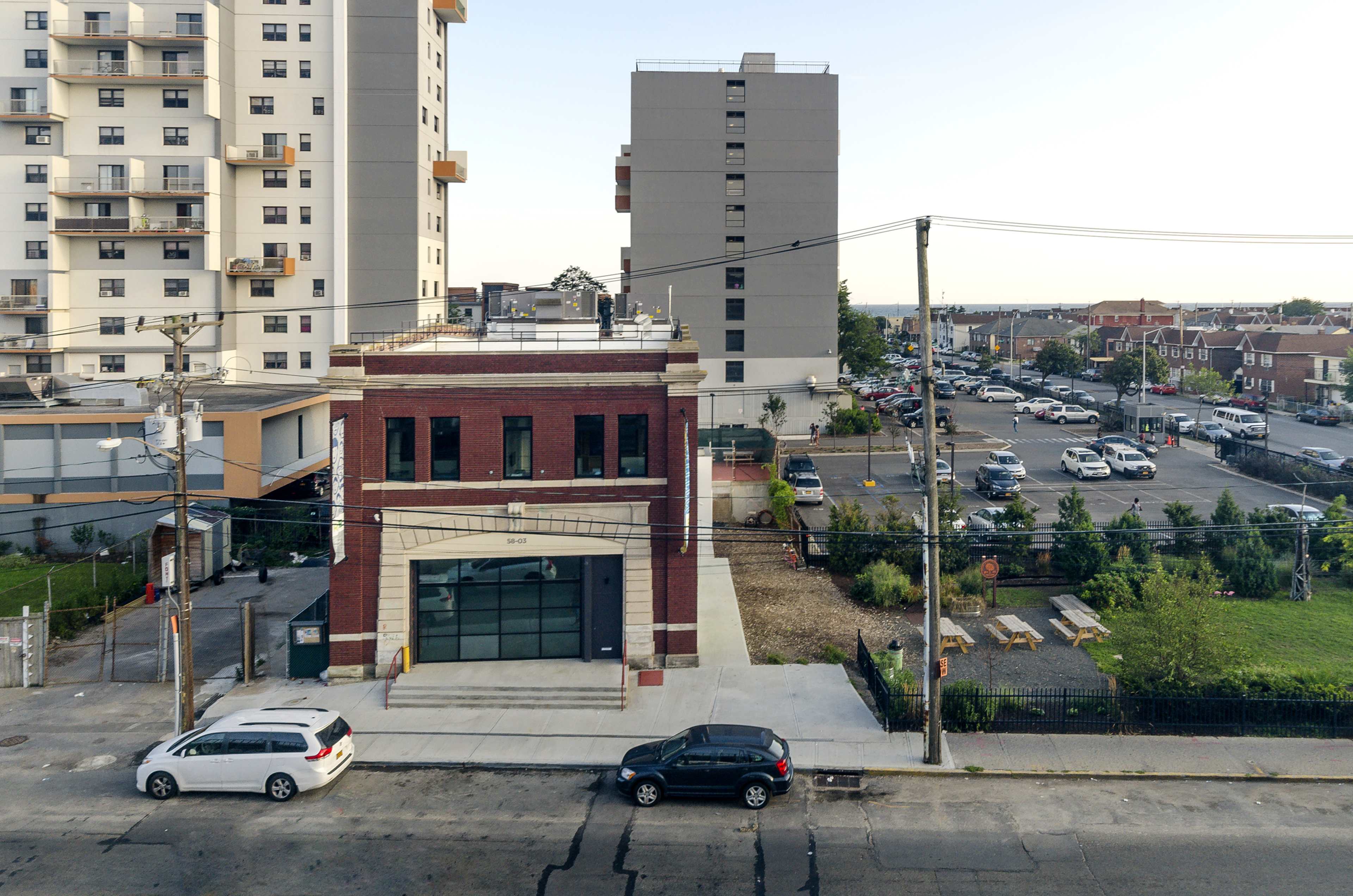 A newly constructed brick building next to taller apartment complexes, with a parking lot and greenery in the background.
