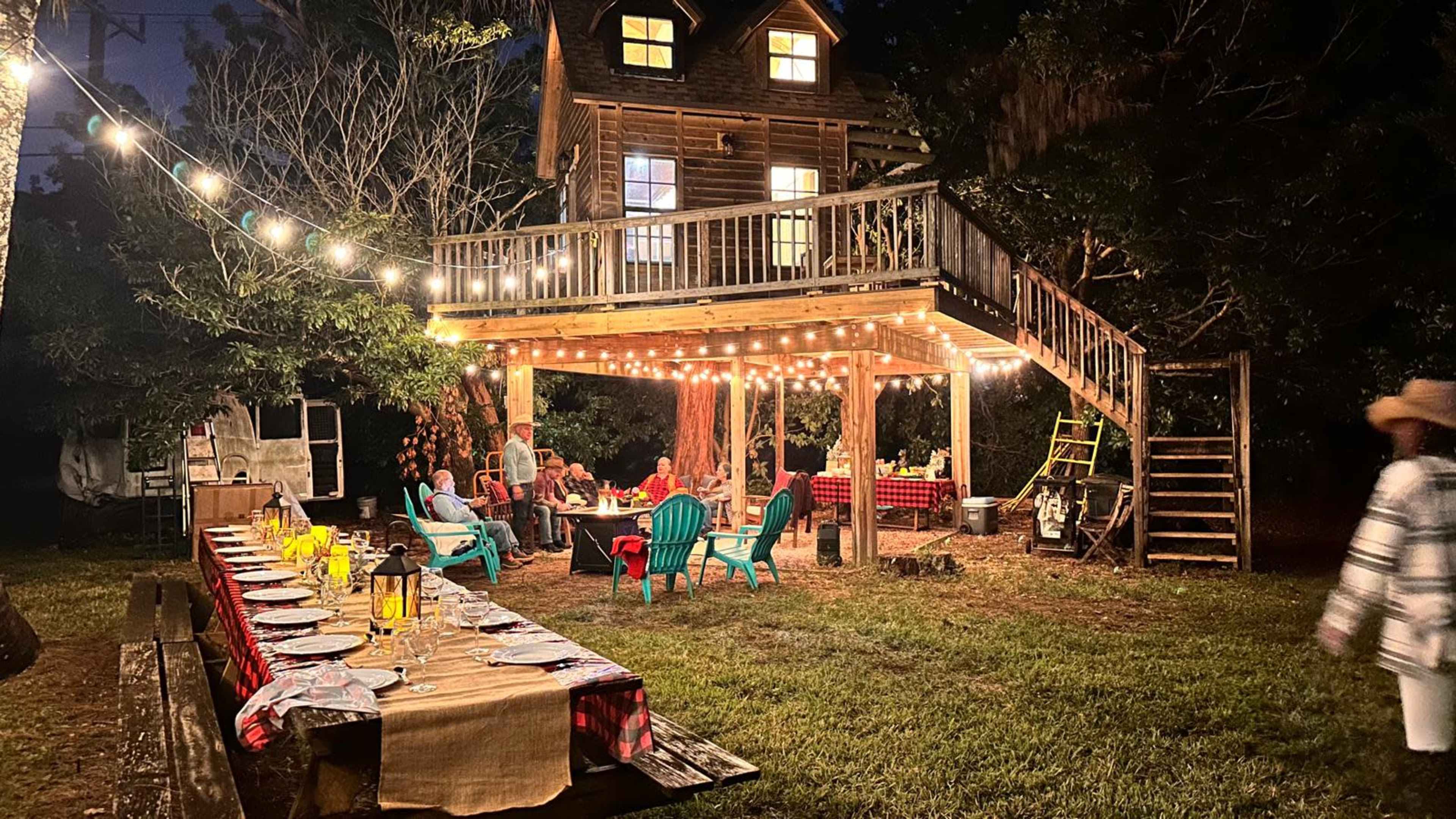 A rustic outdoor dining setup is illuminated by string lights beneath a wooden structure with a playhouse or retreat above it.