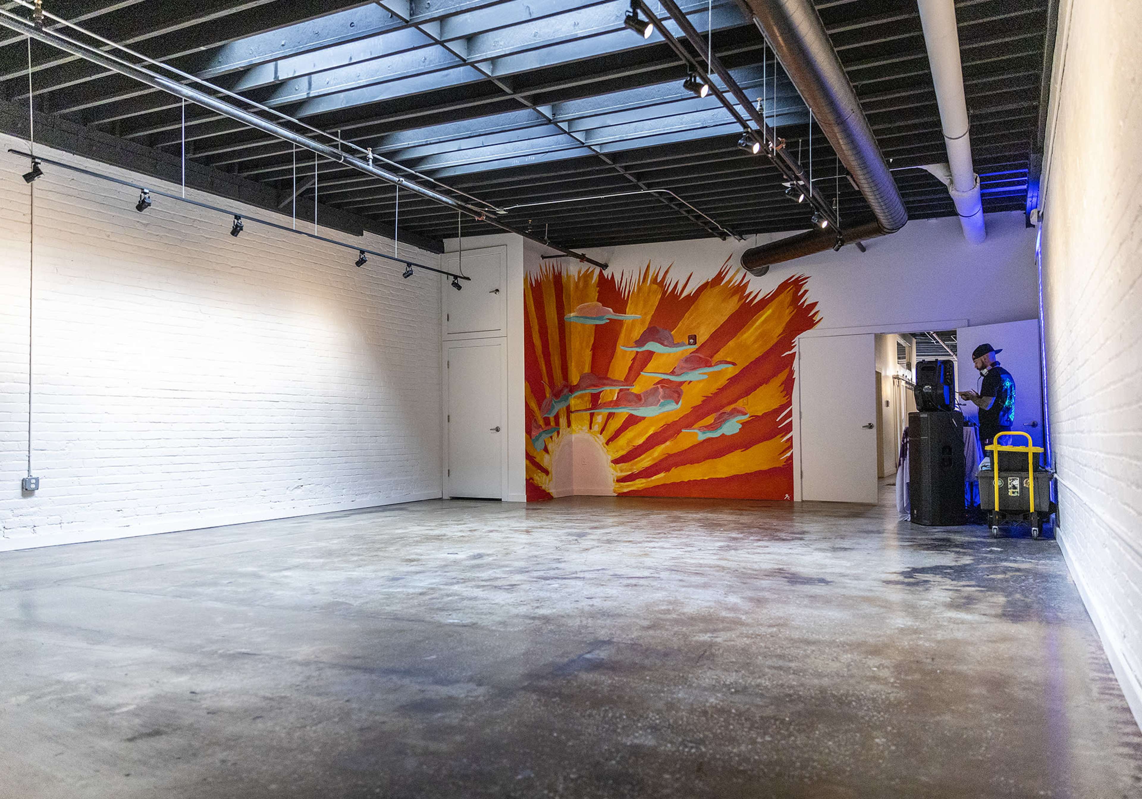 An empty interior space with white brick walls, a painted mural of a sunburst on one wall, and a person standing near a cart in the corner.