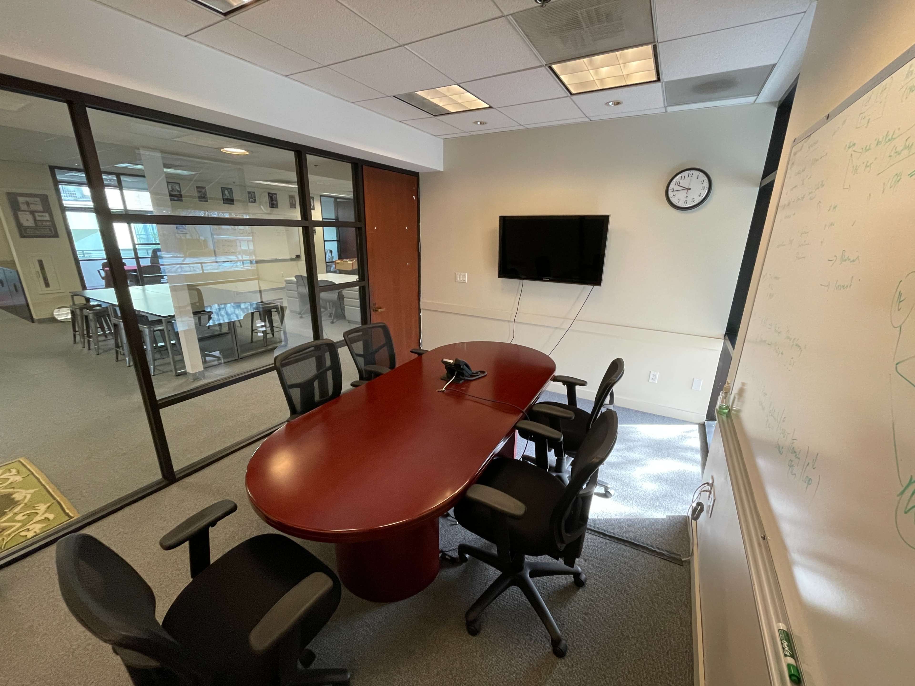 A conference room features a large oval table surrounded by black chairs, a wall-mounted television, and a whiteboard.