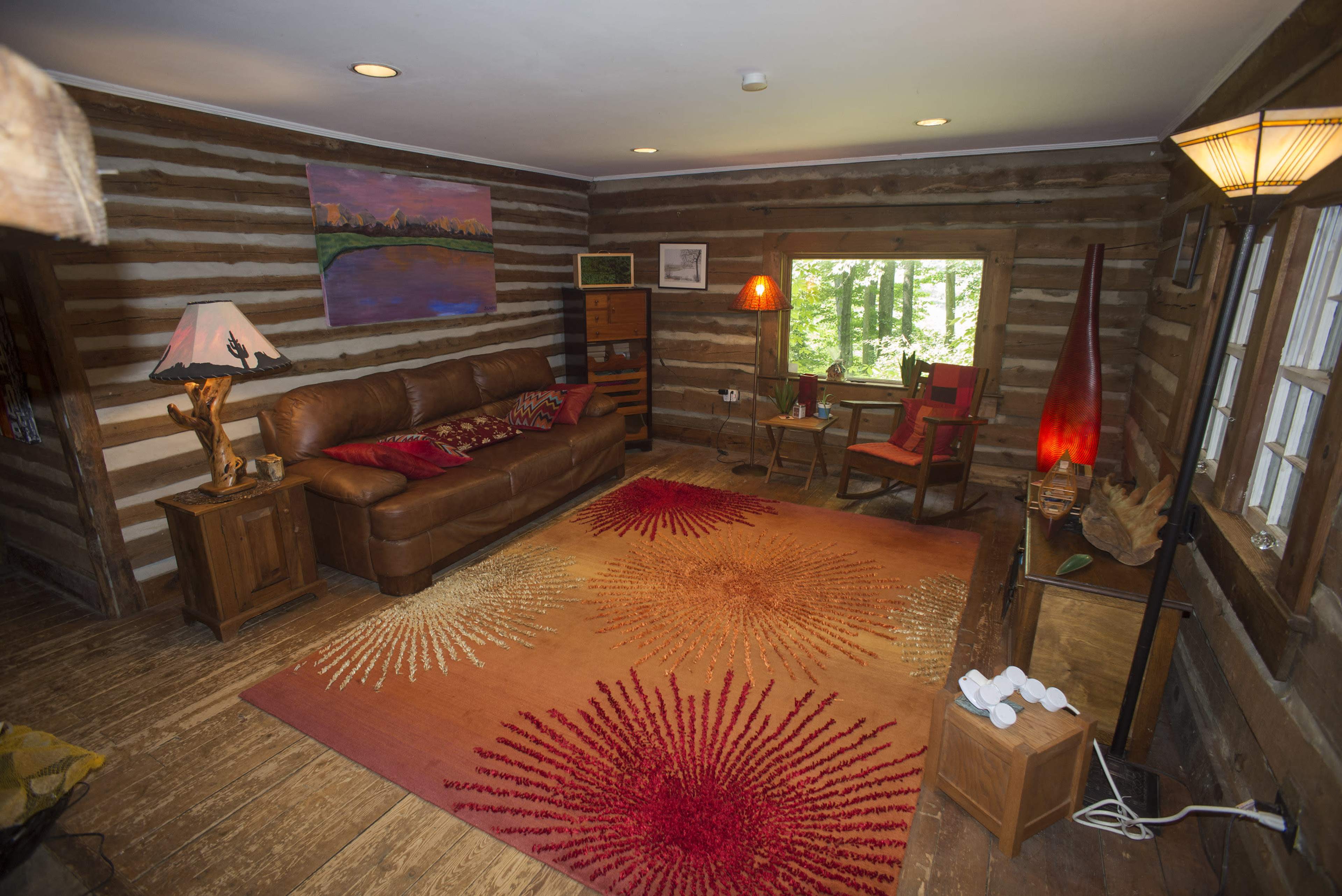 A rustic cabin interior with a leather sofa, wooden furniture, and a bright area rug, surrounded by log walls and a window overlooking trees.
