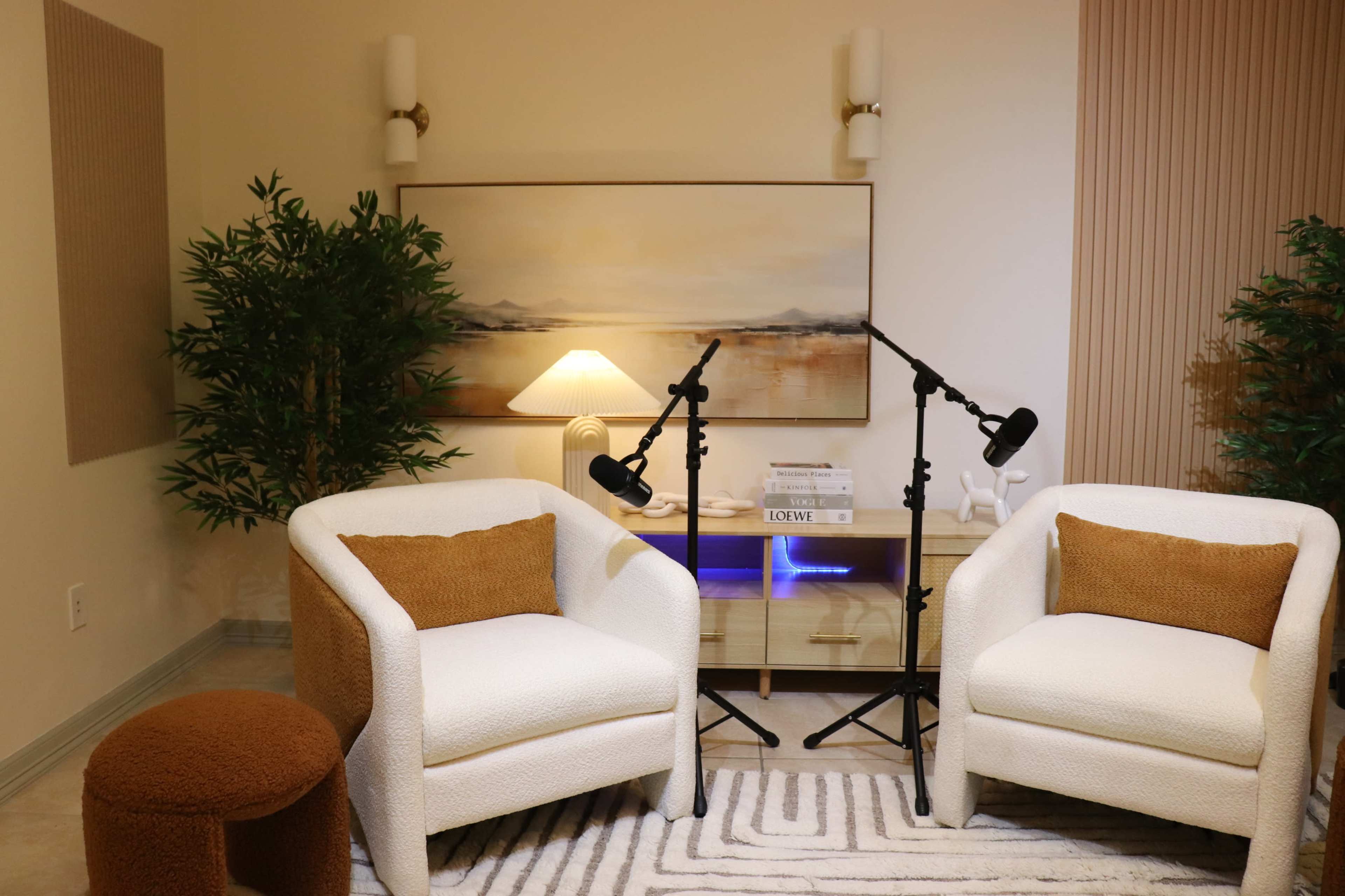 The image shows a cozy room featuring two white upholstered chairs, a small brown stool, a wooden cabinet with blue lighting, and a large landscape painting on the wall, accompanied by two floor lamps and decorative plants.