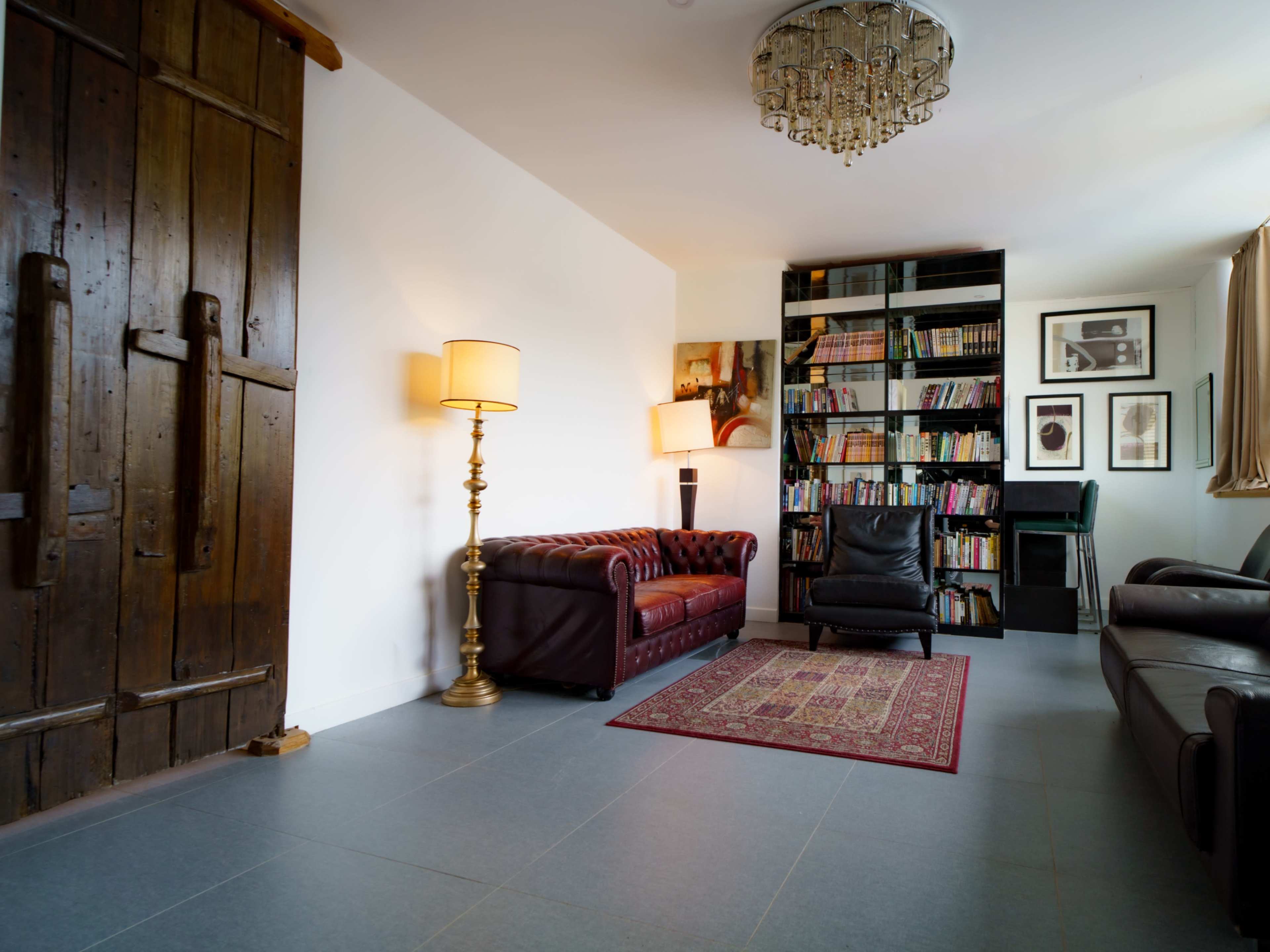 The image shows a spacious living room featuring a brown leather couch, a black armchair, a tall lamp, a bookshelf filled with books, and a wooden door.