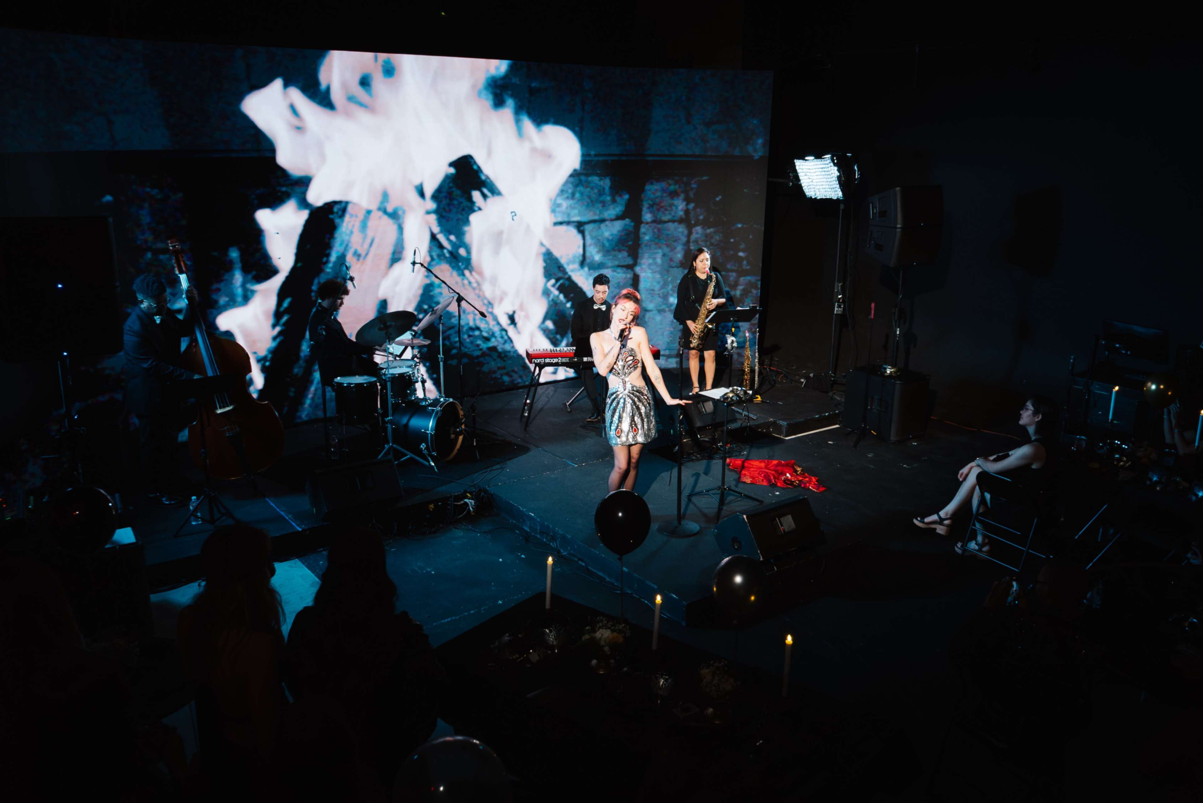 A performer stands on stage in front of a large screen displaying flames, accompanied by a band and an audience seated nearby.
