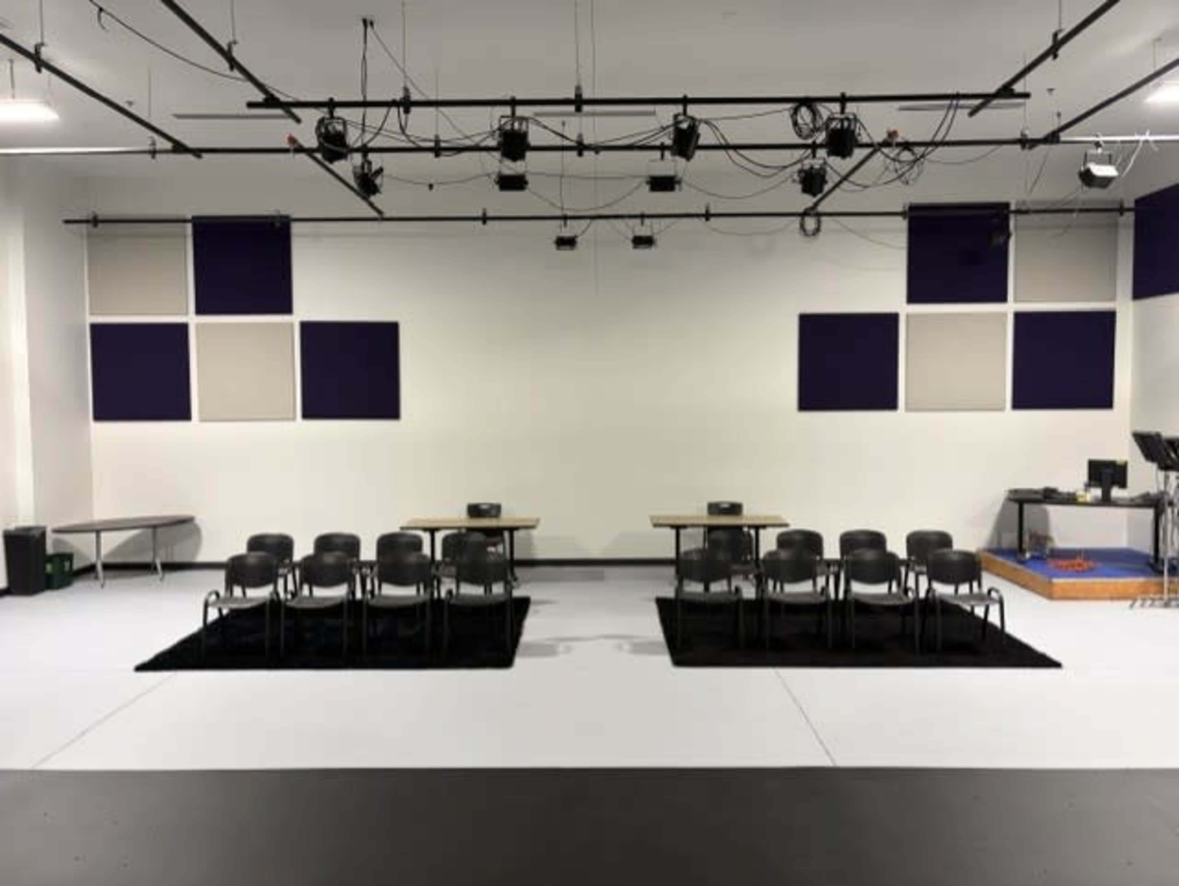 The image shows an empty room with two sets of chairs arranged on black rugs, facing two tables under a ceiling with lighting and audio equipment.