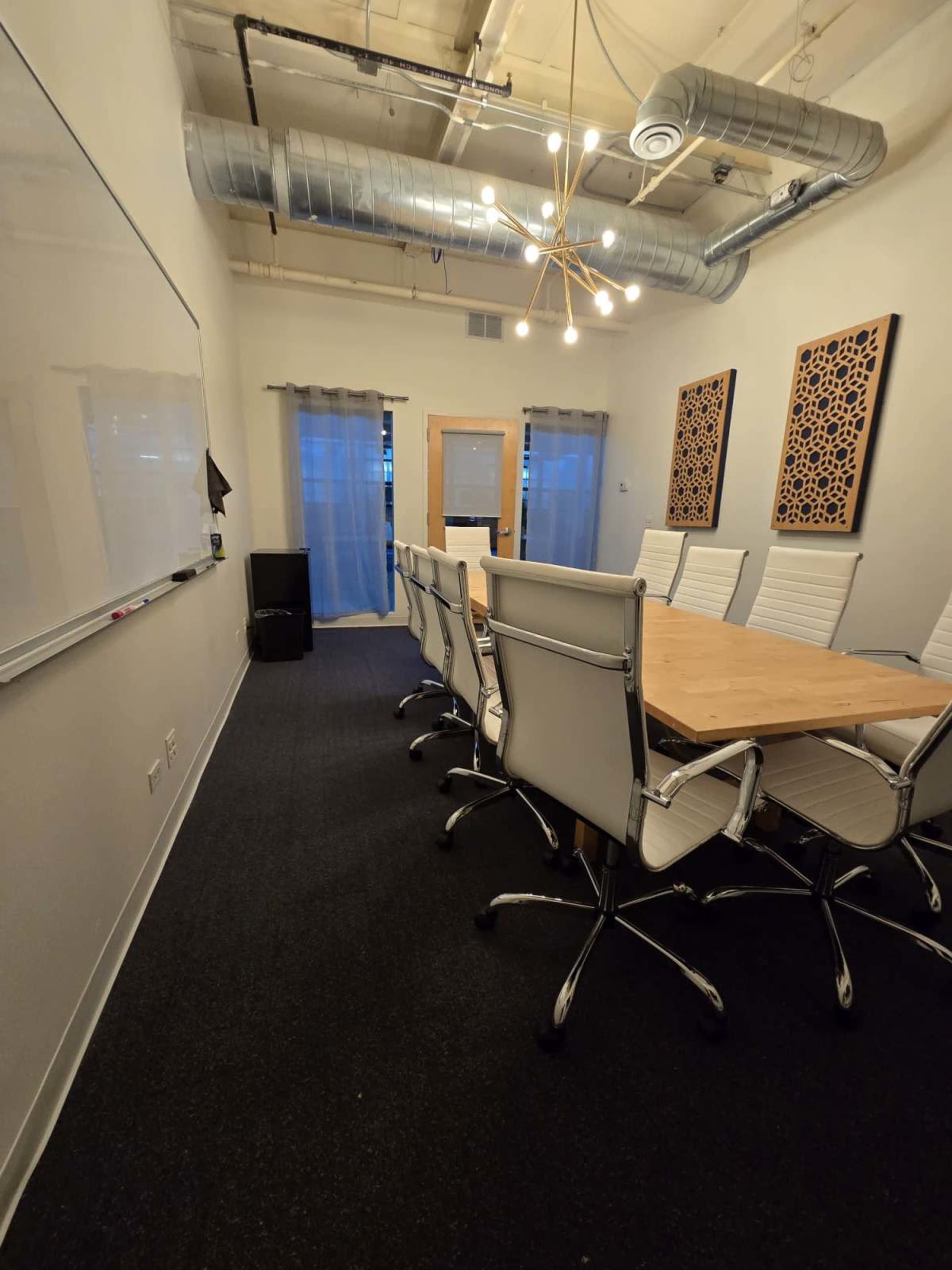 A conference room with a long wooden table surrounded by white chairs, featuring a whiteboard and artwork on the walls.