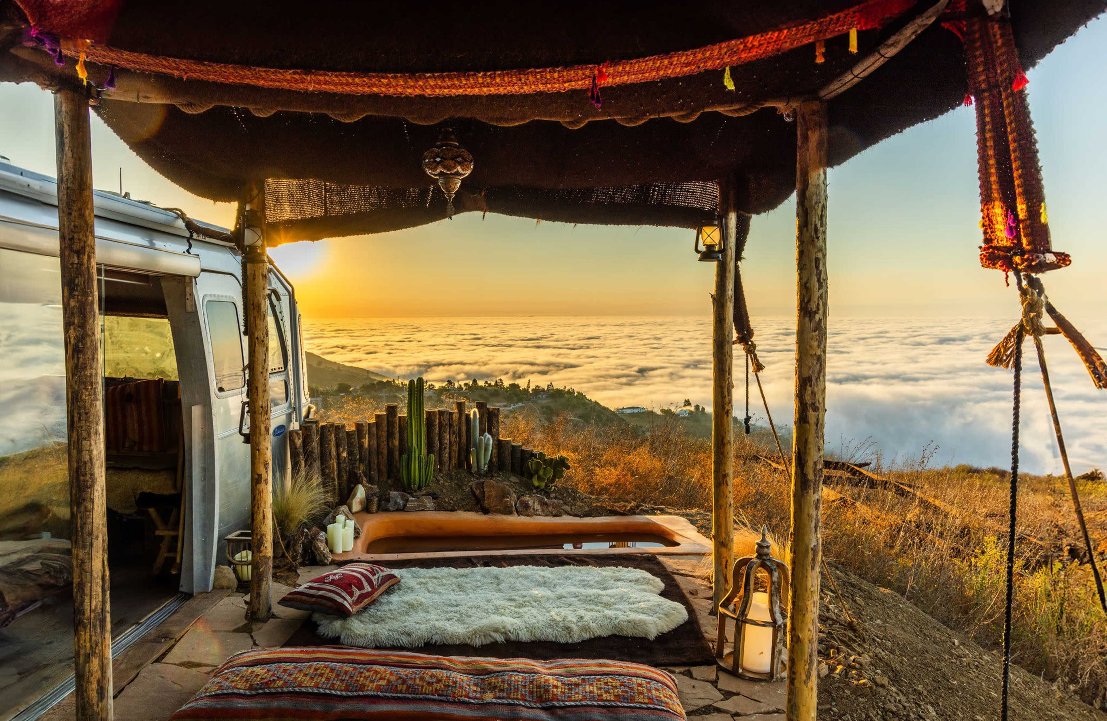 A cozy outdoor space with a plush seating area overlooks a sea of clouds at sunset, framed by rustic wooden posts and a colorful canopy.