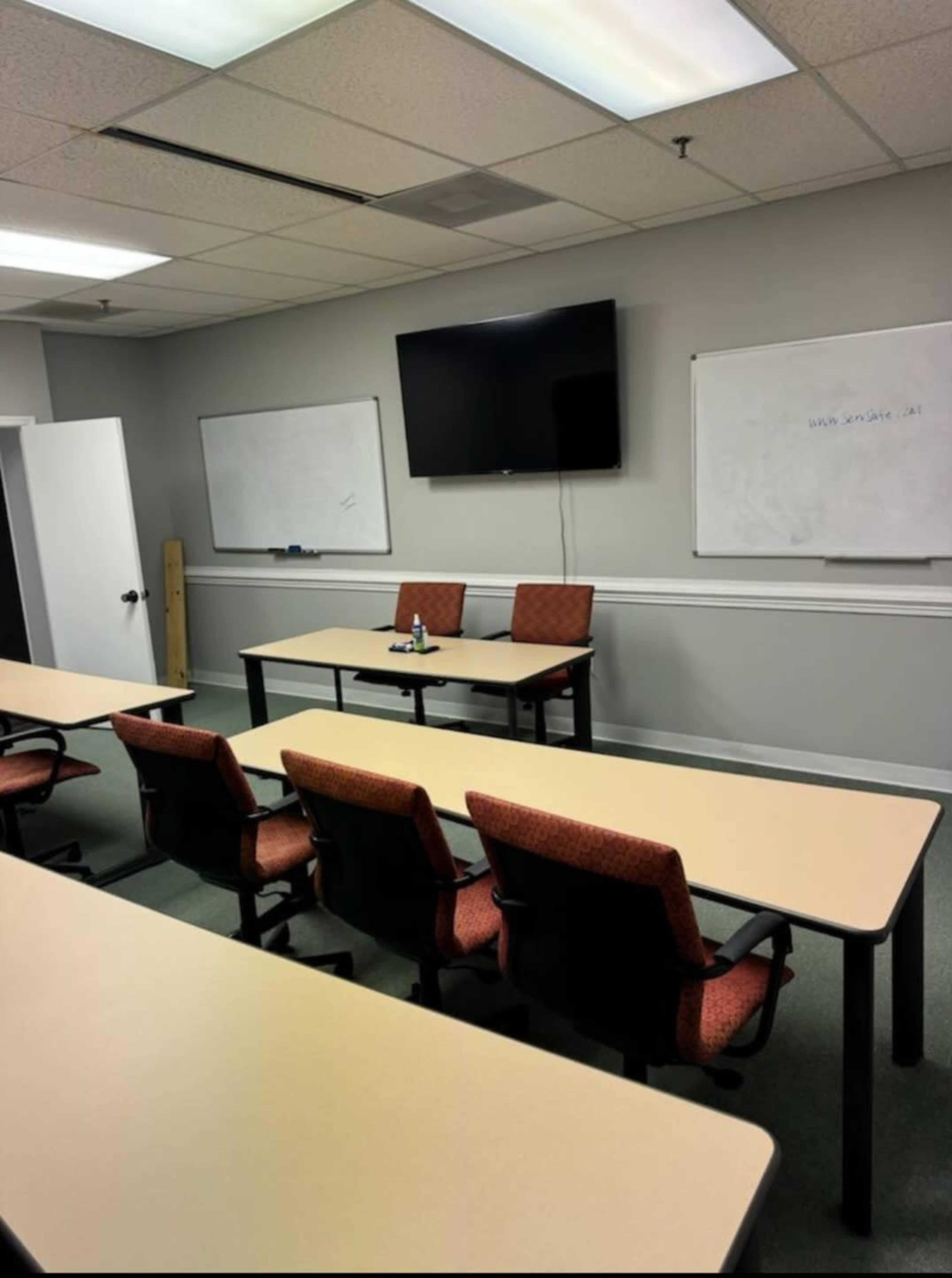 A classroom with rows of chairs and tables facing a television mounted on the wall, and two empty chairs at the front.