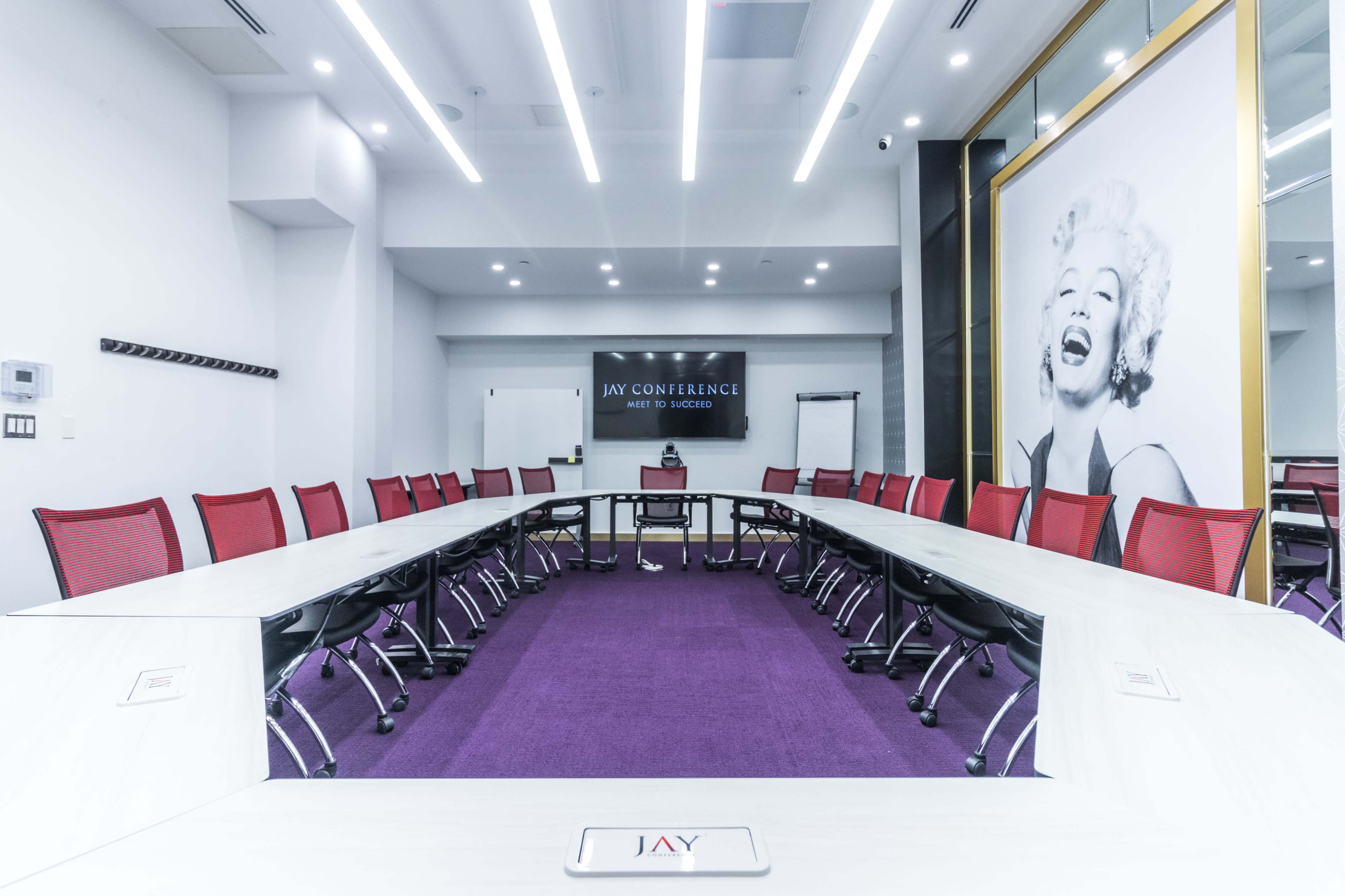 A modern conference room features a large rectangular table surrounded by red chairs, with a purple carpet and a prominent portrait of Marilyn Monroe on the wall.