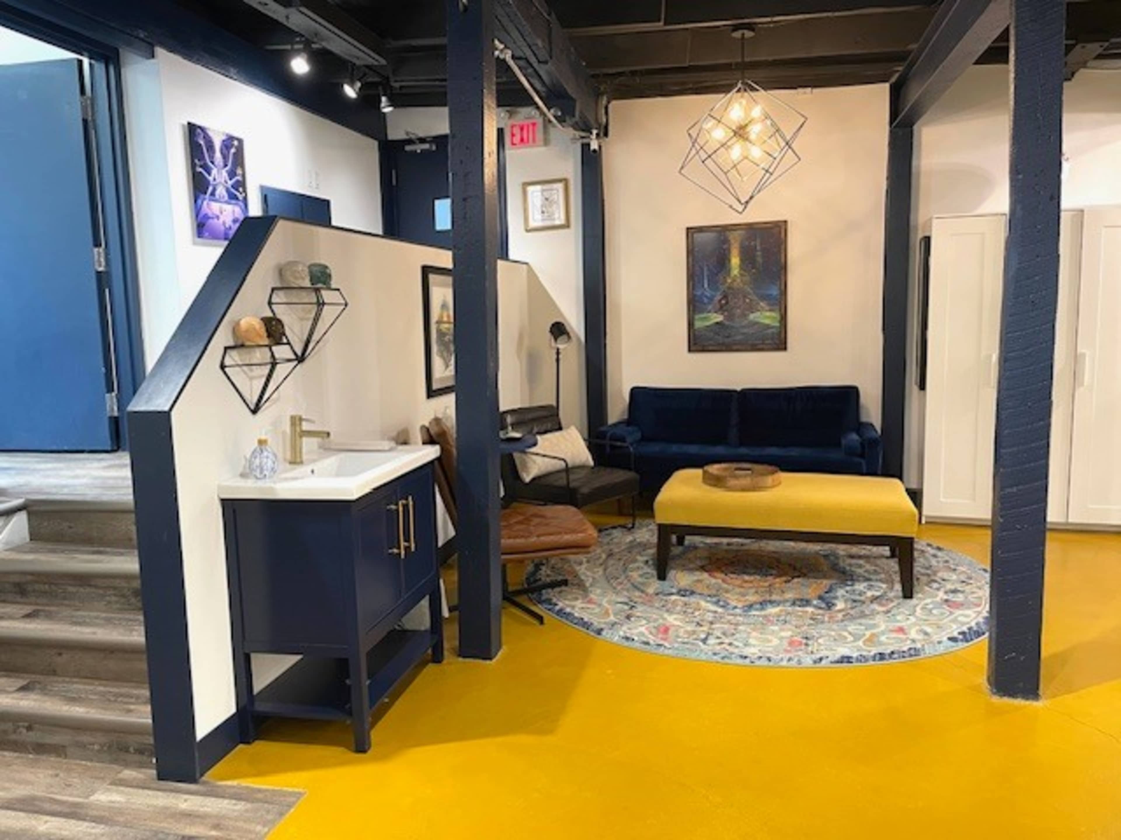 The image shows a modern interior space featuring a blue sofa, a yellow coffee table, and a geometric light fixture, with stairs leading to another level.