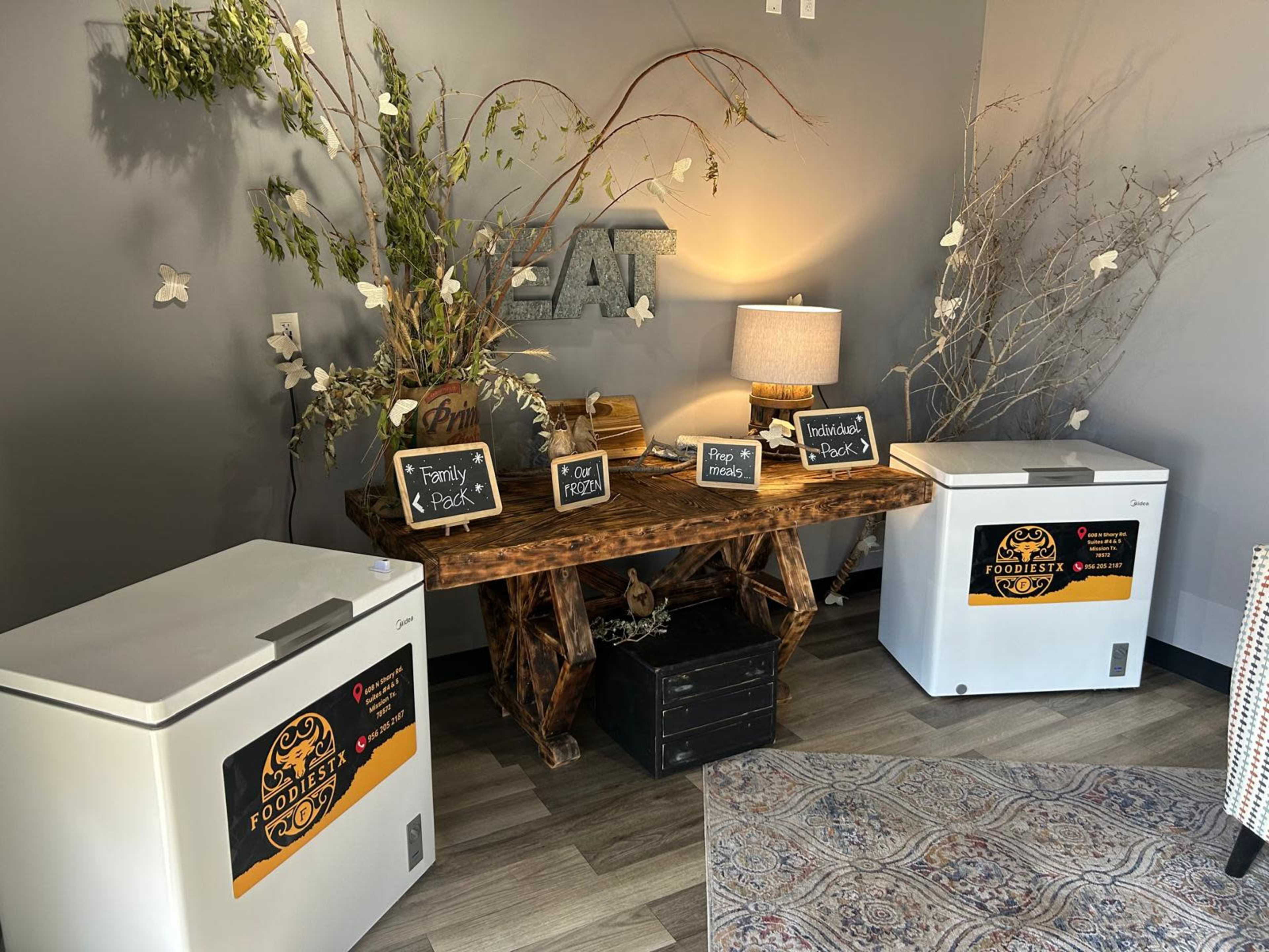 The image shows a small corner of a room featuring a wooden table with decorative elements, two white freezers labeled with signage, a lamp, and some greenery.