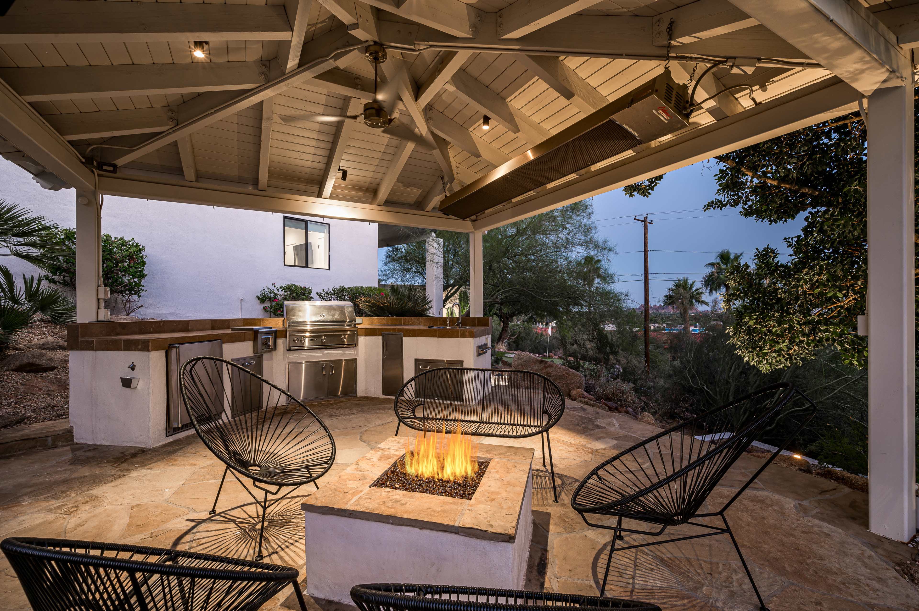 An outdoor patio features a fire pit surrounded by stylish chairs and an adjacent built-in grill area under a covered roof.