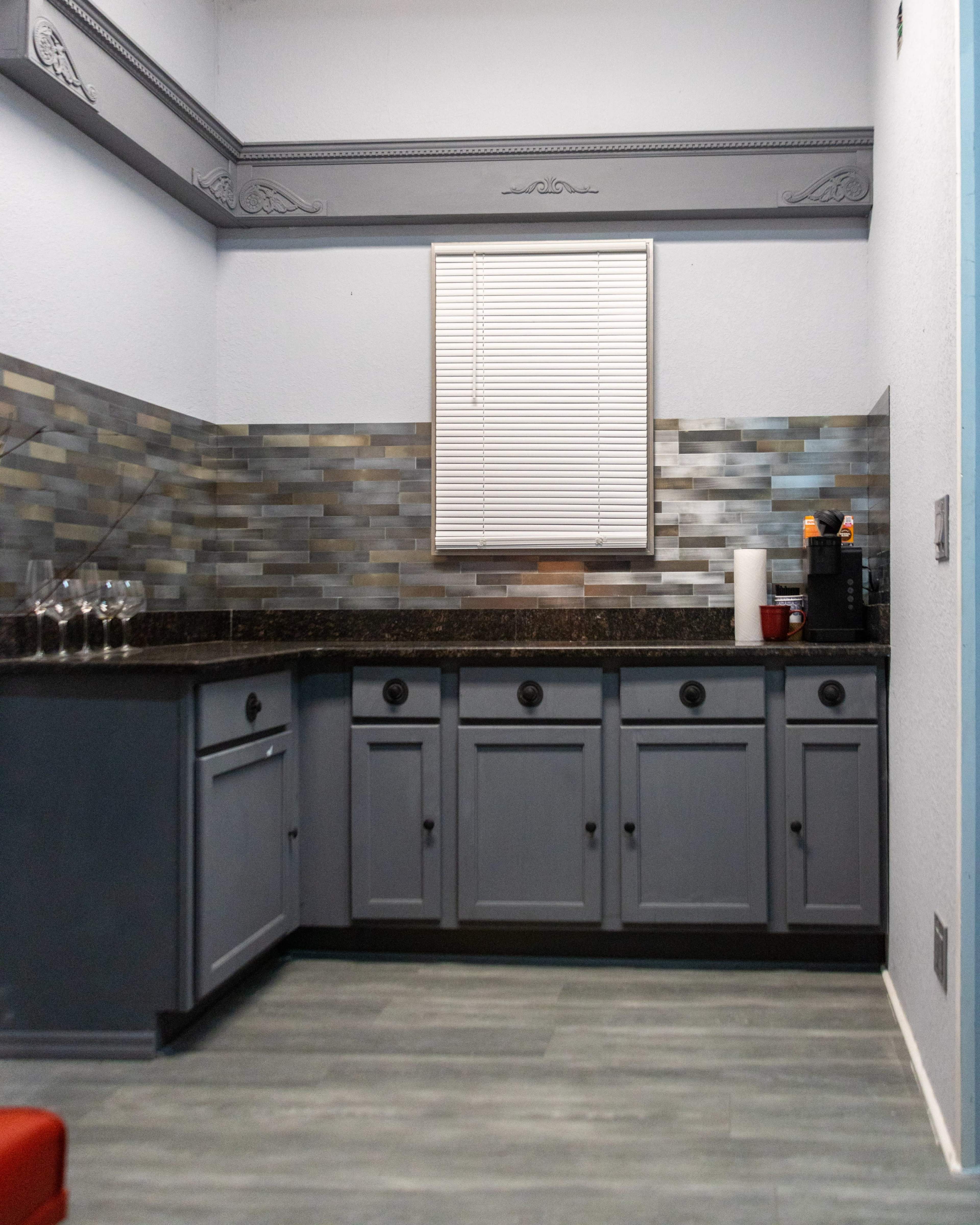 The image shows a kitchen with a gray cabinet and countertop, a tiled backsplash, and a window covered by a white blind.