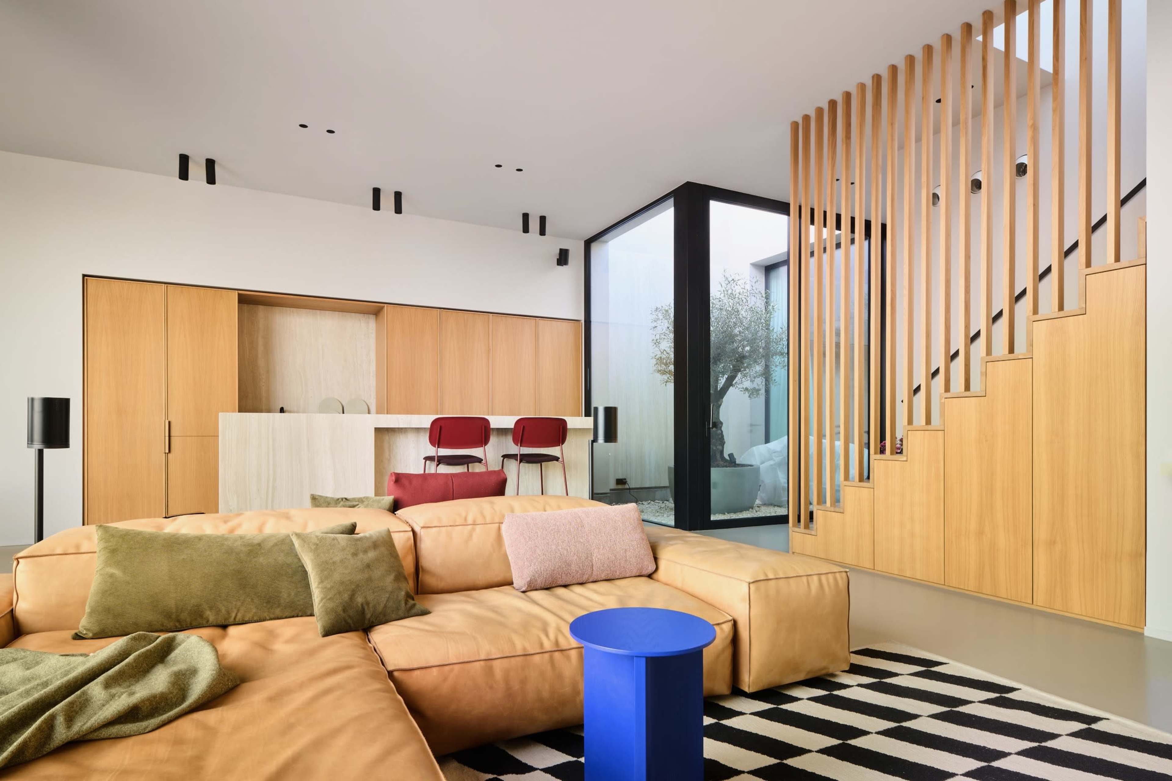 The image shows a modern living room featuring a large, tan sectional sofa, a blue side table, a staircase with wooden slats, and a minimalist kitchenette with red bar stools.
