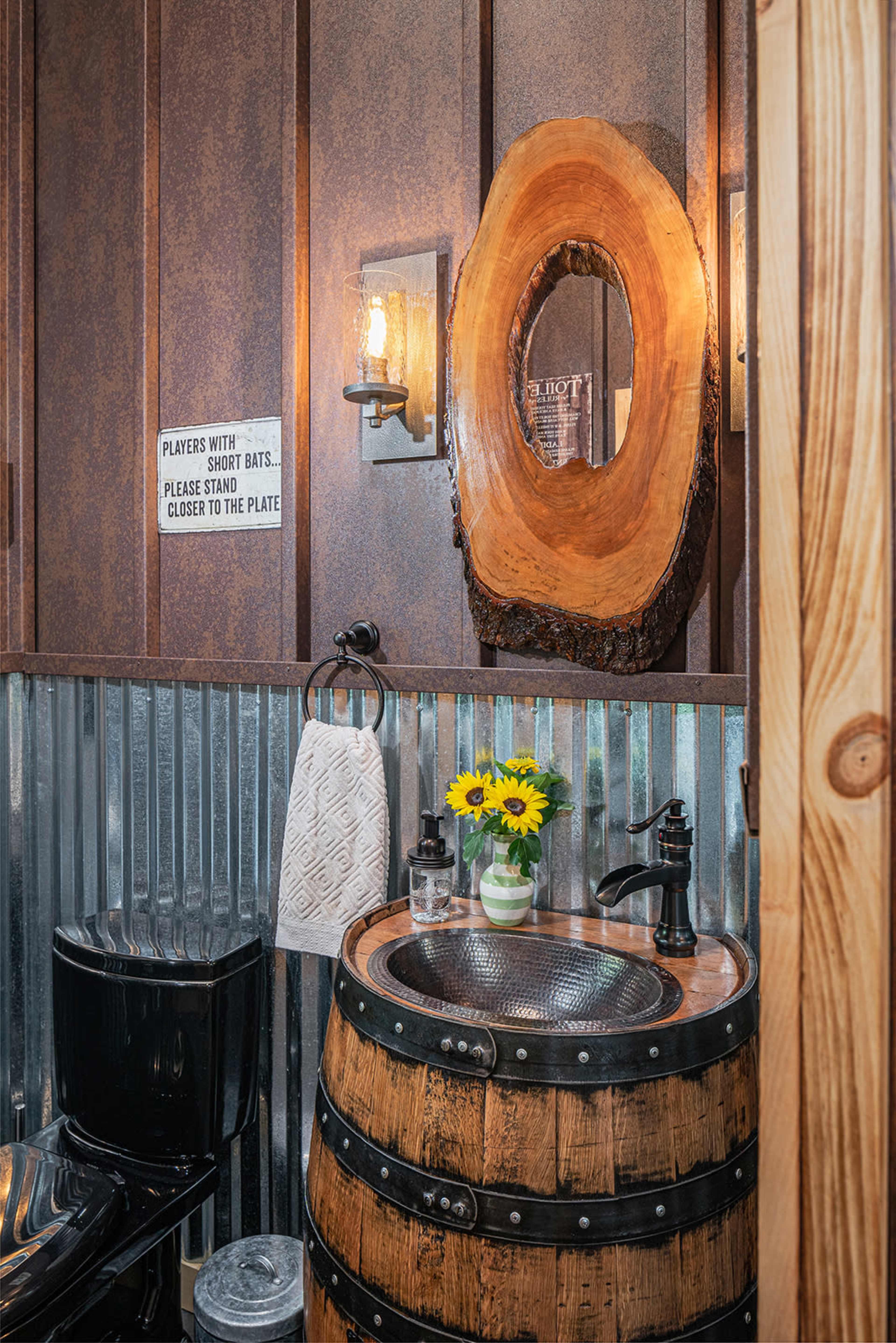 A rustic bathroom featuring a barrel sink, a wooden mirror, and a wall with metal sheeting and a floral arrangement.