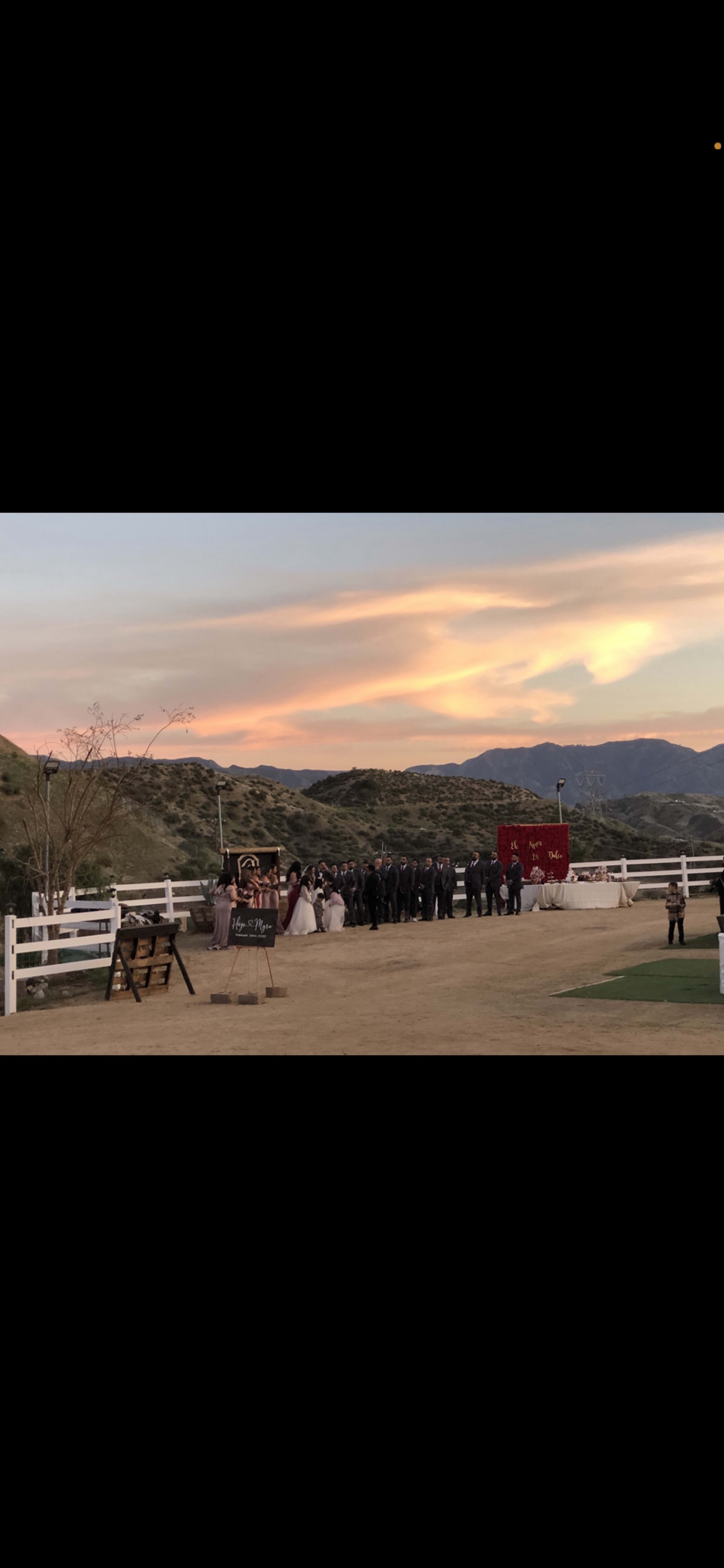 A wedding ceremony is taking place outdoors with a group of people in formal attire standing in front of a bride and groom, against a backdrop of mountains and a colorful sunset.
