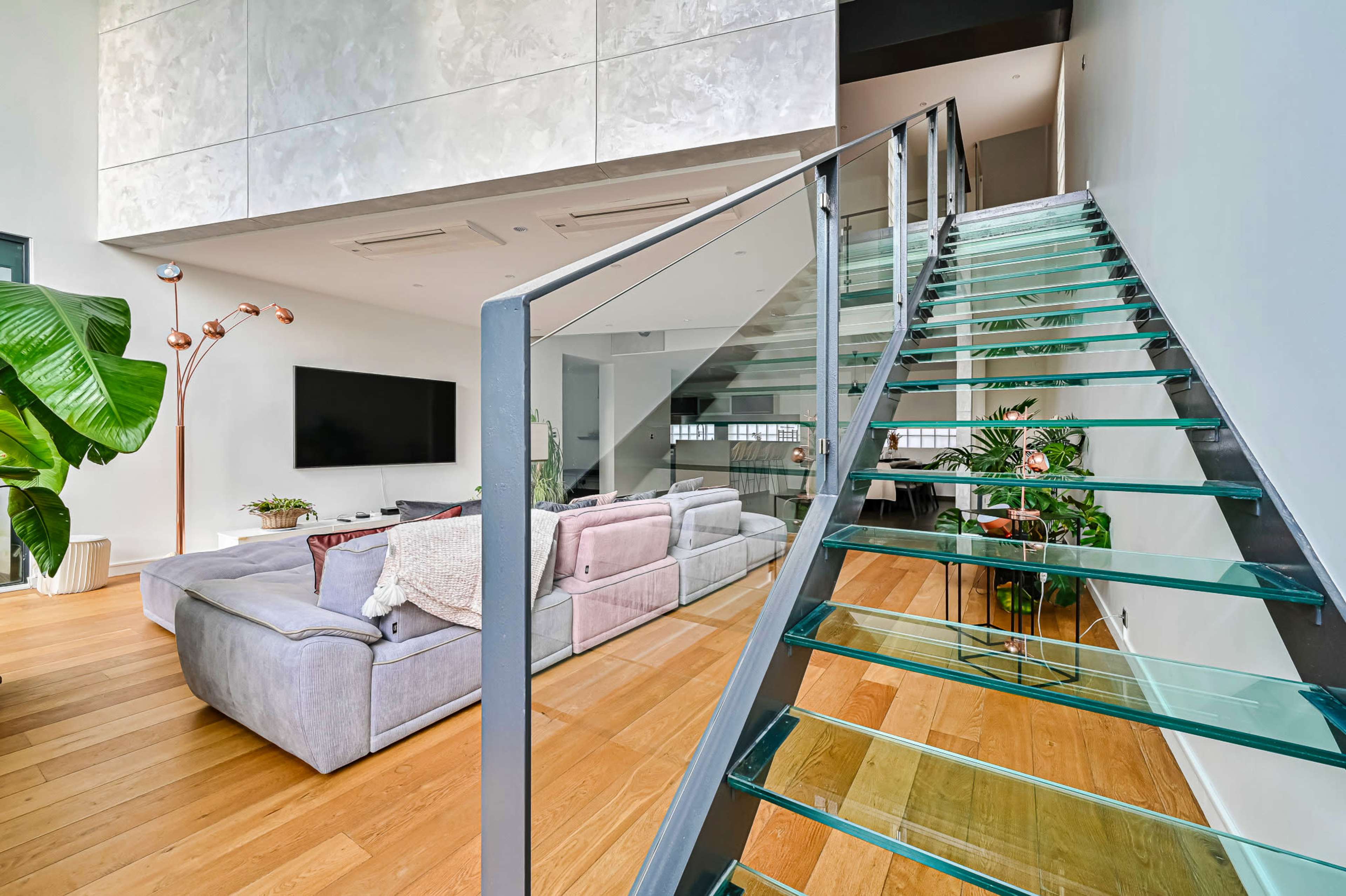 A modern interior features a glass staircase leading to an upper level, with a spacious living area that includes a large sofa and a television.