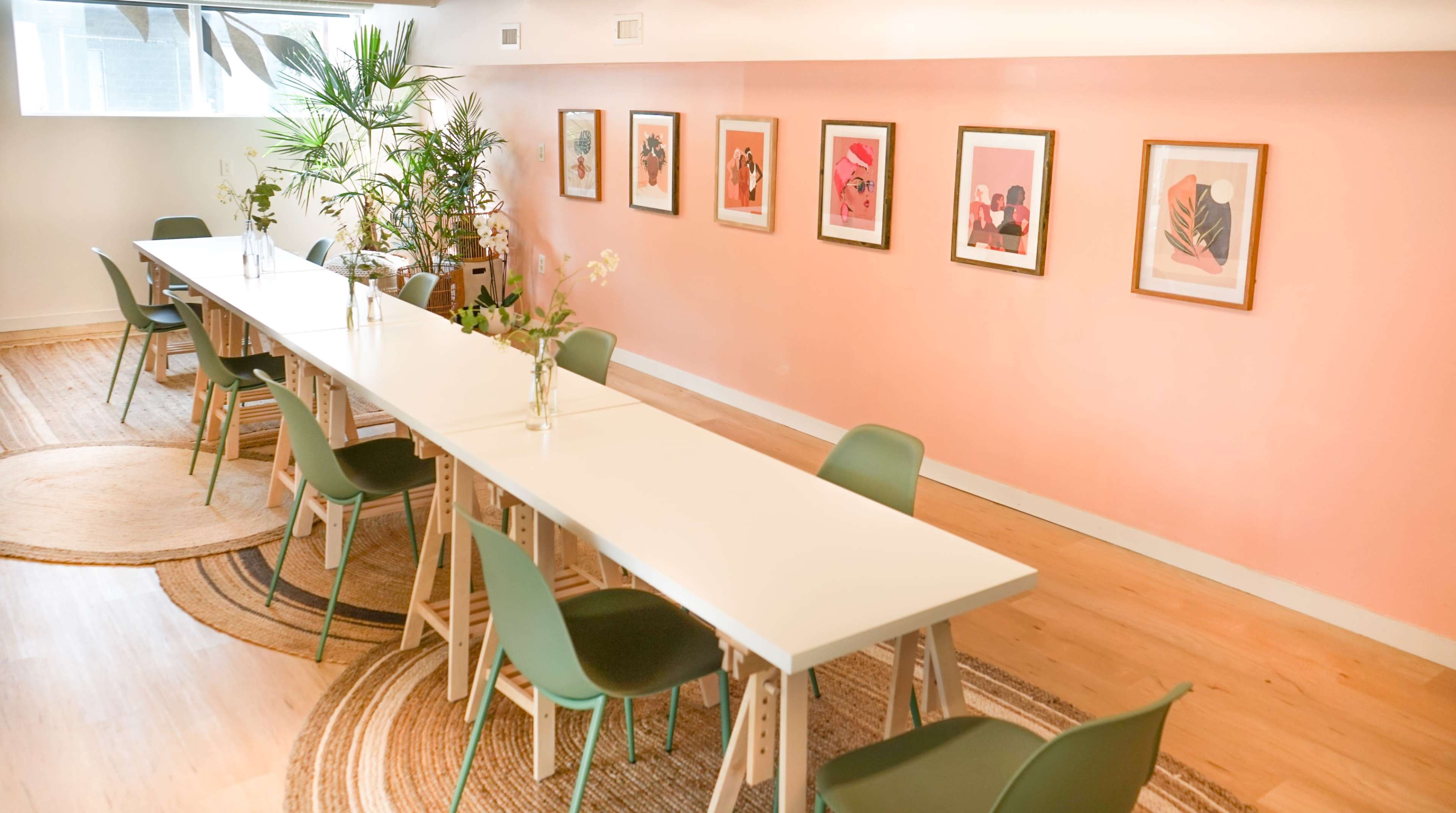 A long, white table with green chairs is set in a room featuring a pink wall adorned with framed artwork and plants.
