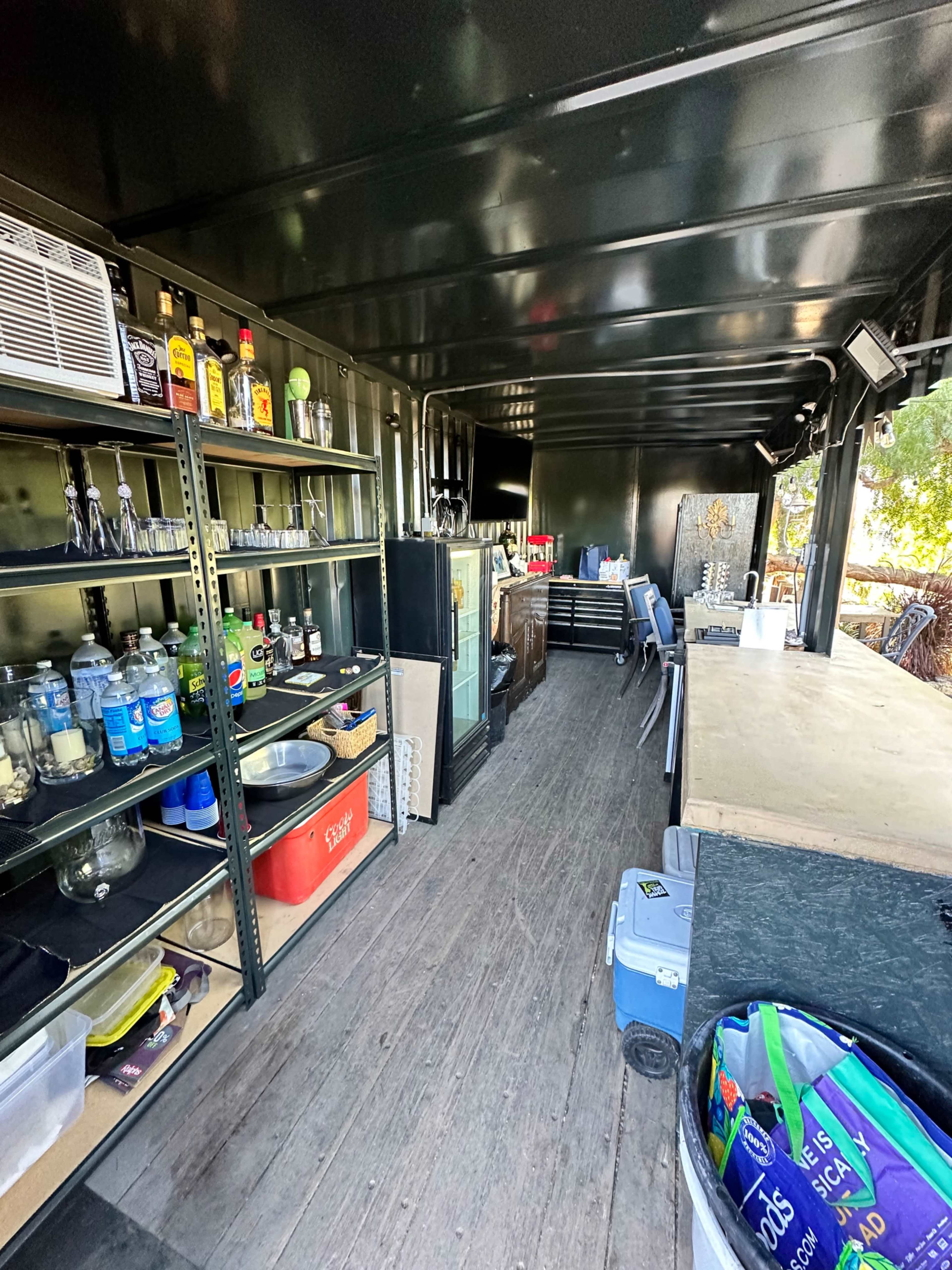 The interior of a commercial kitchen or bar setup inside a shipping container, featuring shelves stocked with beverages and utensils, a large countertop, and kitchen appliances.