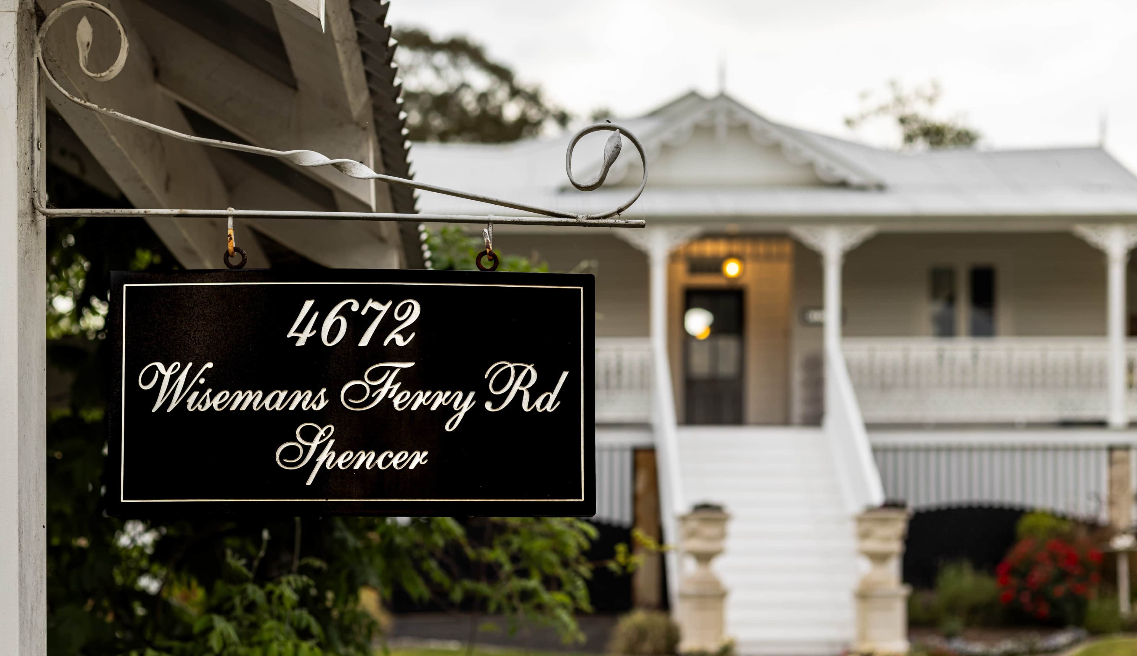 A black sign displays the address "4672 Wisemans Ferry Rd" in front of a two-story house with a white facade and steps leading up to a porch.