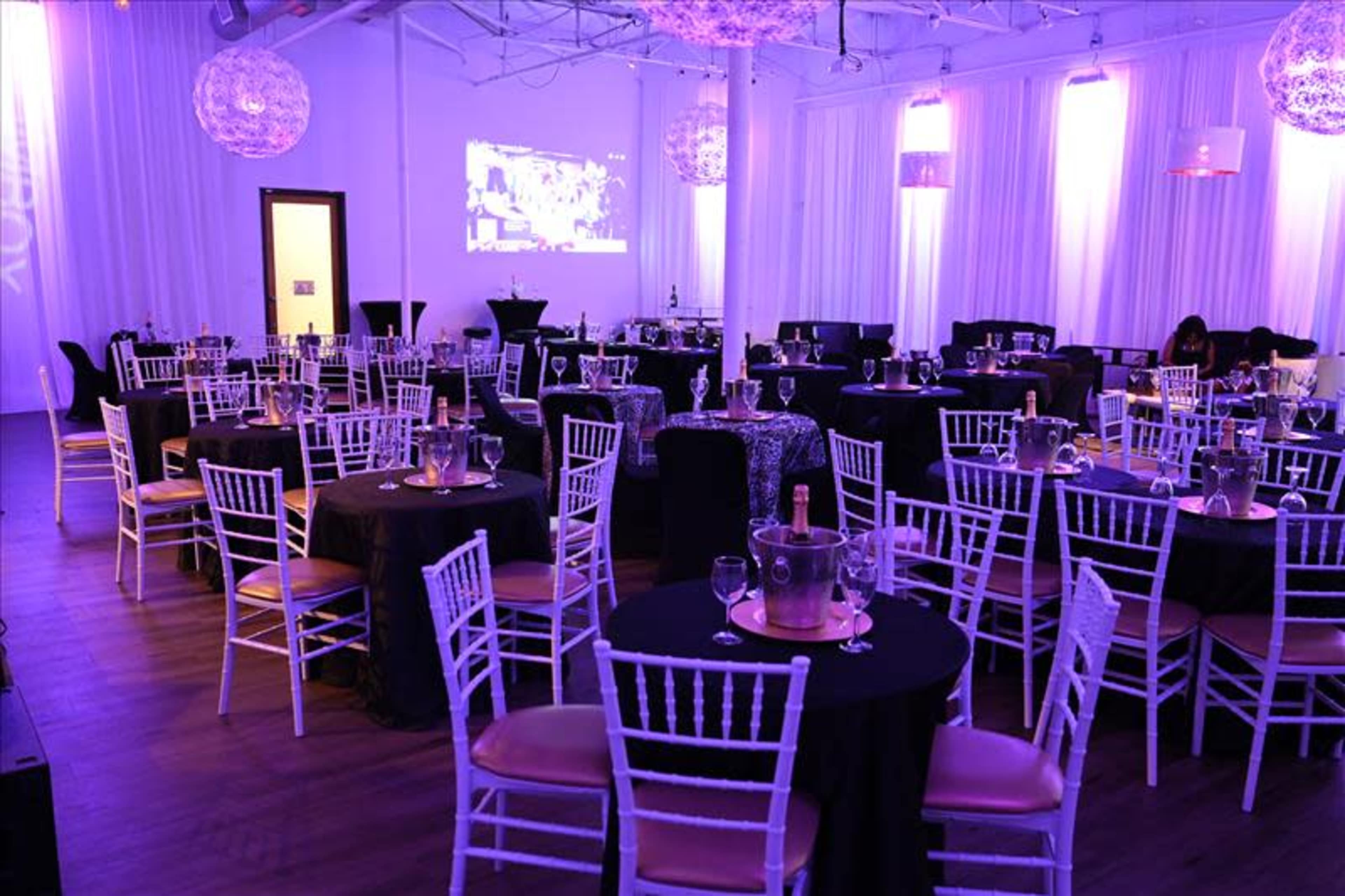 A well-decorated event space with round tables set for guests, featuring white chairs, black tablecloths, and ambient lighting.