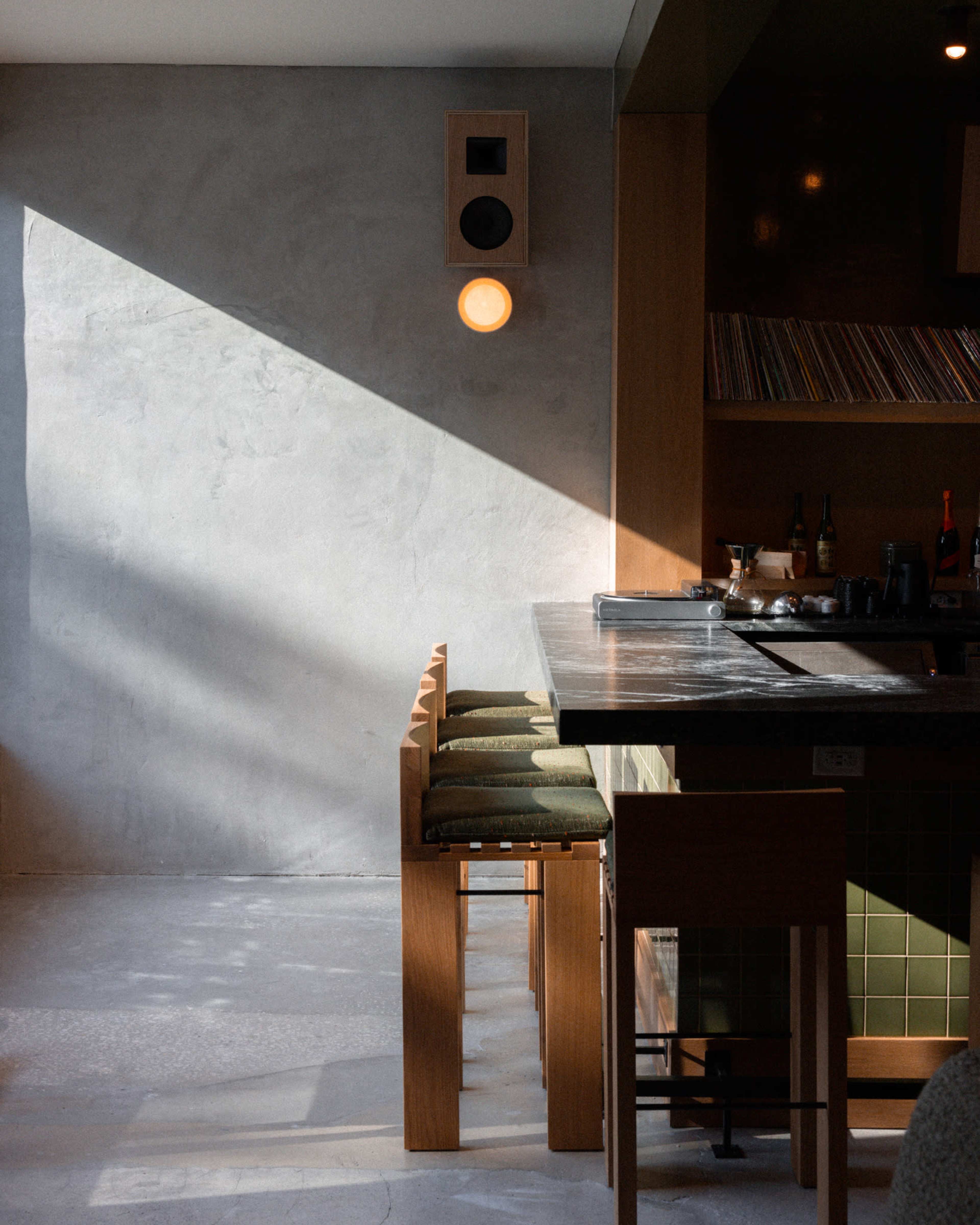A minimalist cafe interior features a wooden counter with high stools, a concrete wall, and a warm light fixture casting shadows.