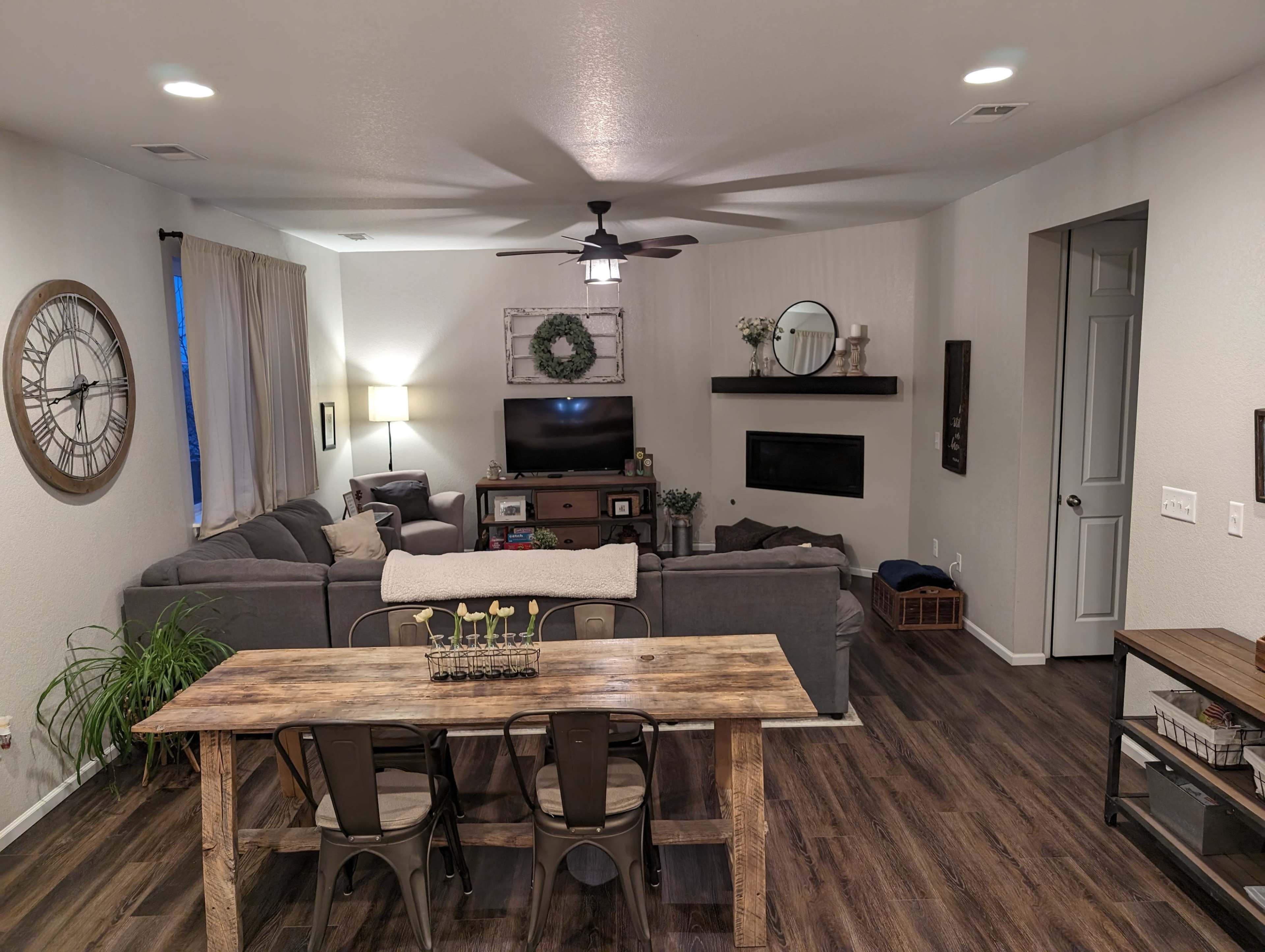 A modern living room with a gray sectional couch, a wooden dining table with metal chairs, a television mounted on the wall, and decorative elements like a clock and wreath.
