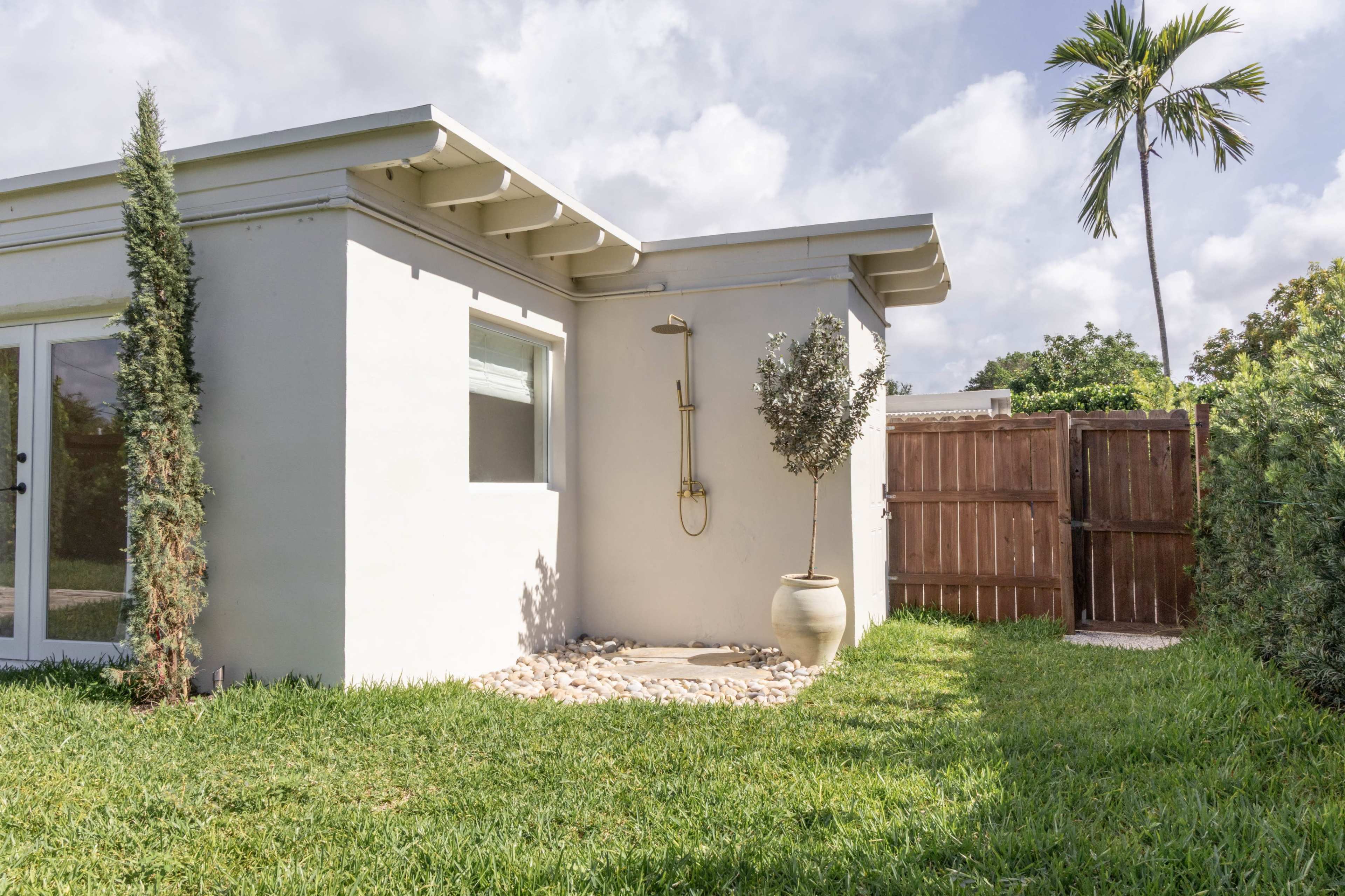 The image shows a modern outdoor shower attached to a white building surrounded by green grass and a wooden fence.
