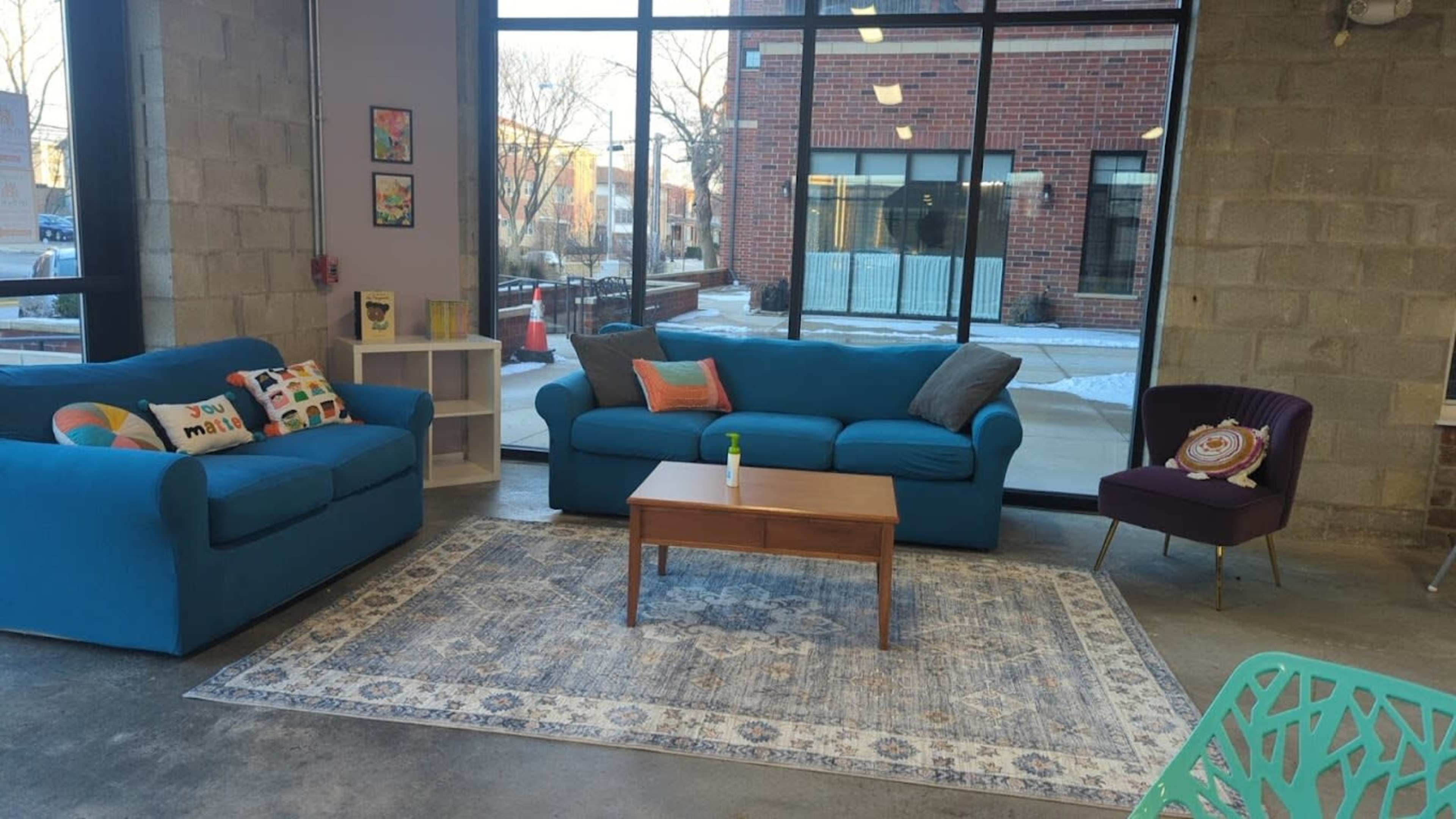 A modern lounge area features two blue sofas, a wooden coffee table, and a patterned rug, framed by large windows showcasing the outdoors.