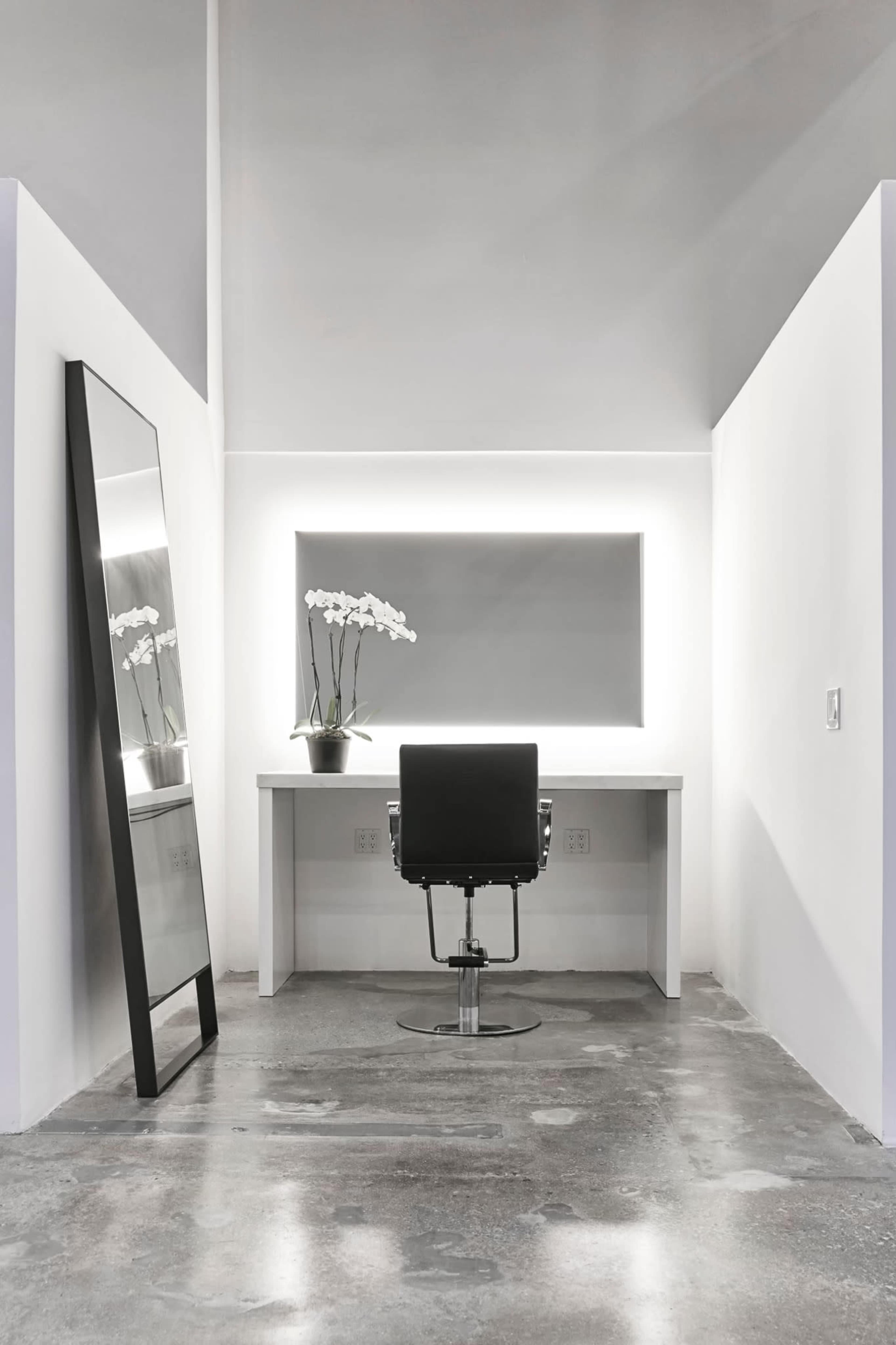 A minimalist workspace featuring a white desk, a black swivel chair, a large mirror, and an orchid plant on the desk, all against a plain wall.