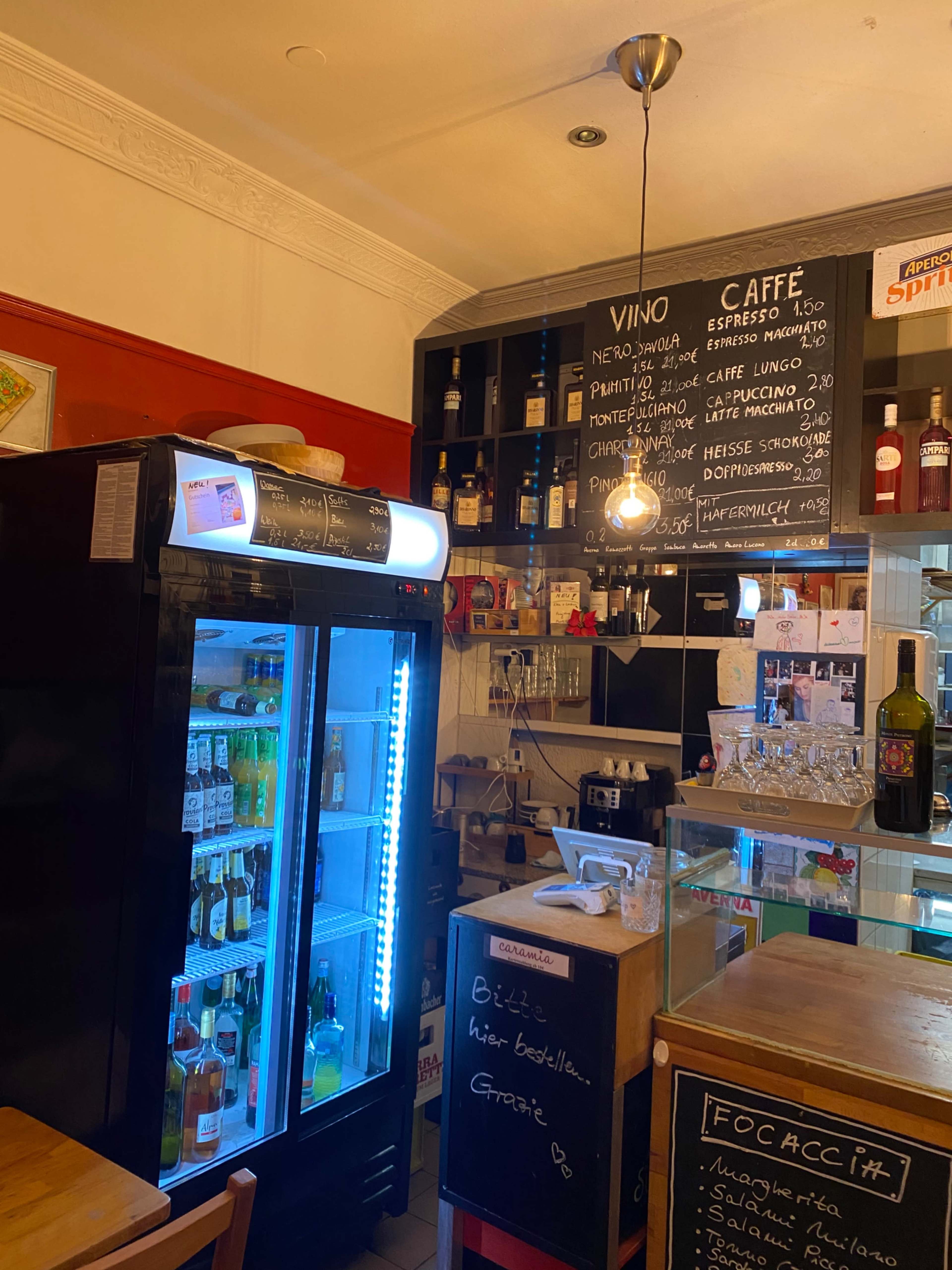 The image shows a small café interior with a glass-fronted refrigerator displaying various beverages, a chalkboard menu, and a wooden counter with a bottle selection.