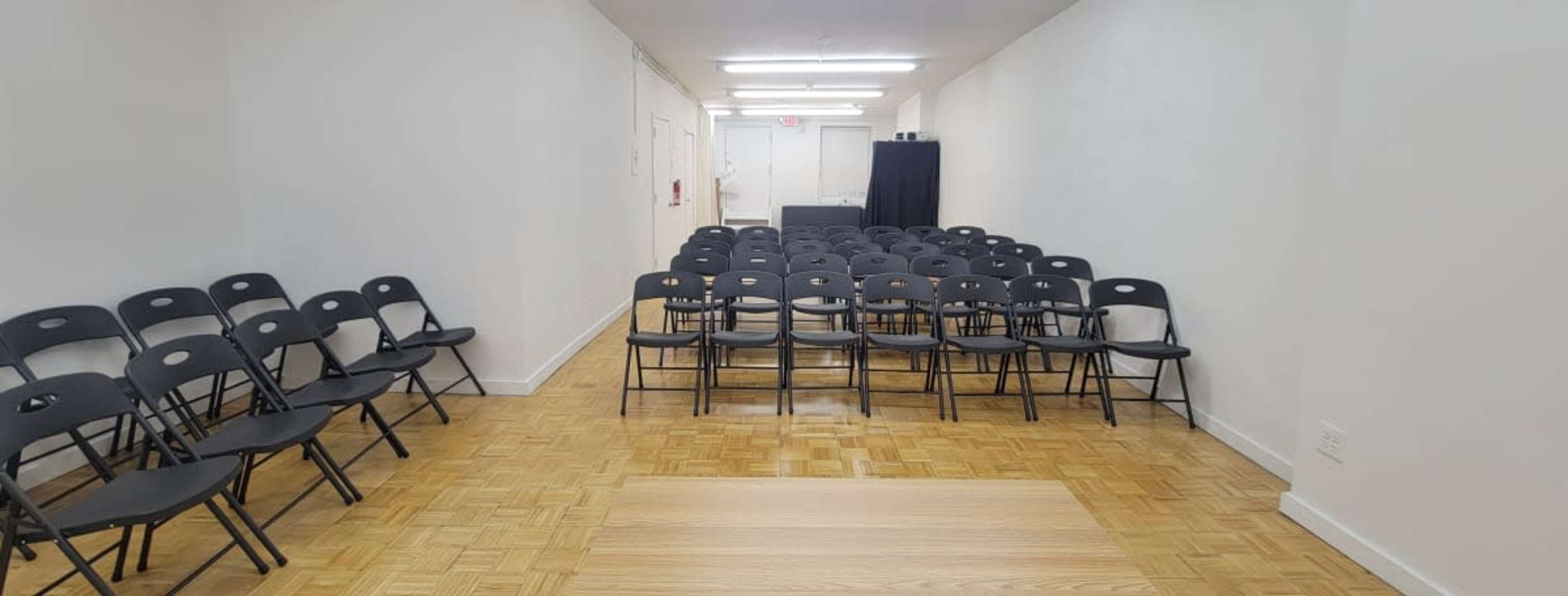 The image shows a long, narrow room with rows of black folding chairs facing a wooden stage or platform at the front.