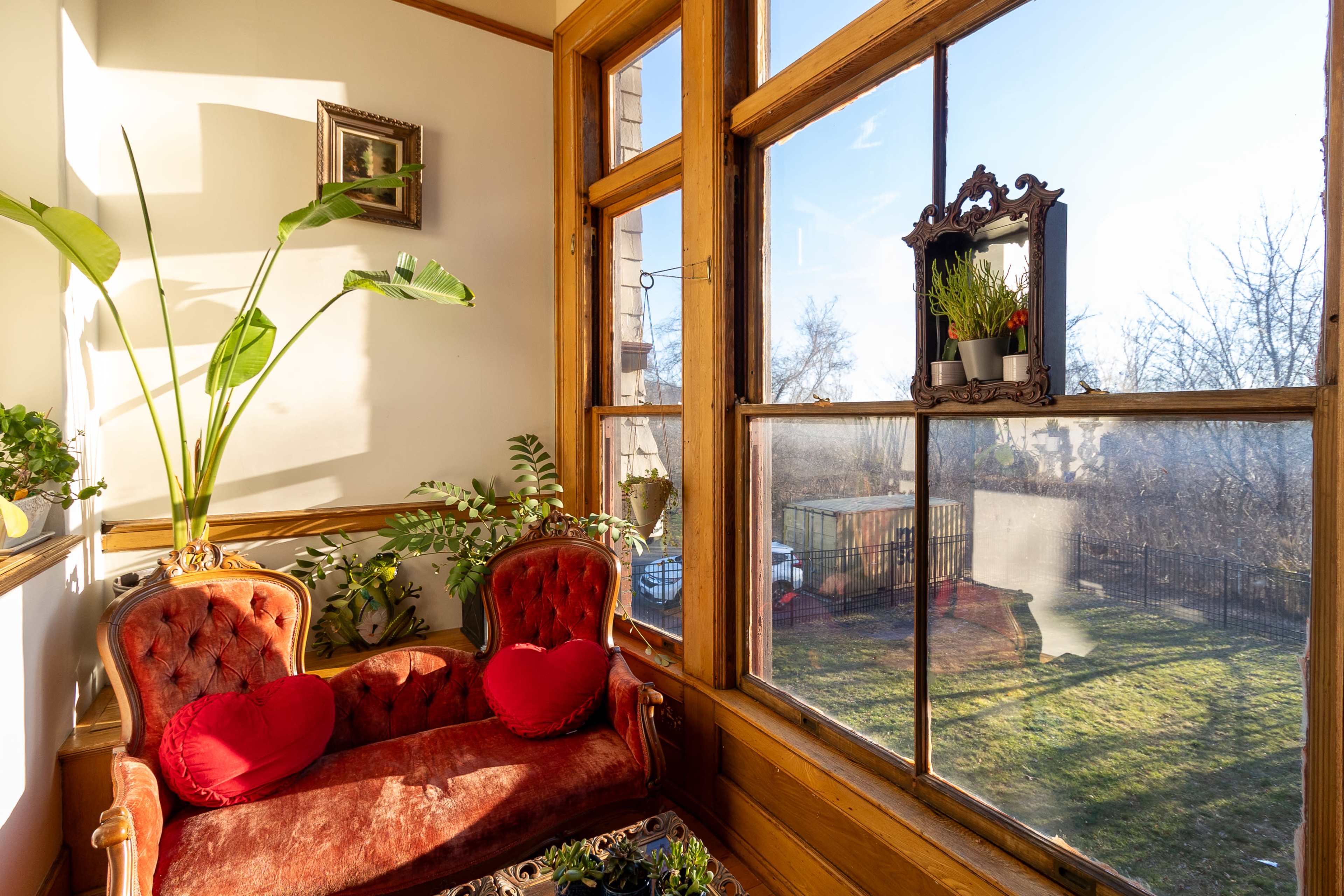 A cozy sunlit corner features a vintage red velvet couch with decorative pillows, surrounded by plants and large windows overlooking a grassy yard.