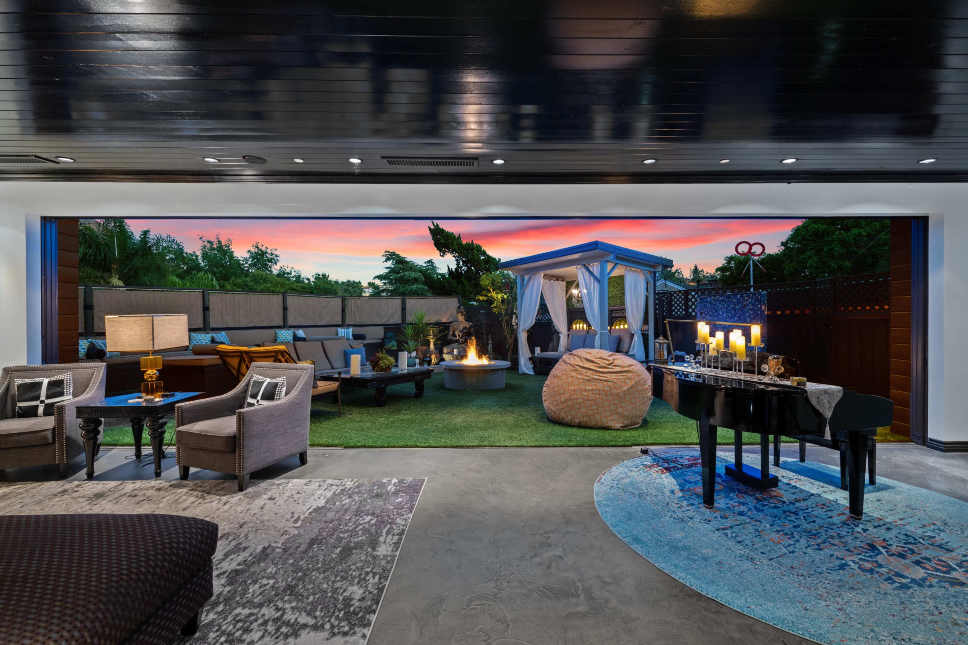 A modern indoor lounge area with a view of an outdoor patio featuring a fire pit, seating, and a spacious lawn, all set against a sunset backdrop.