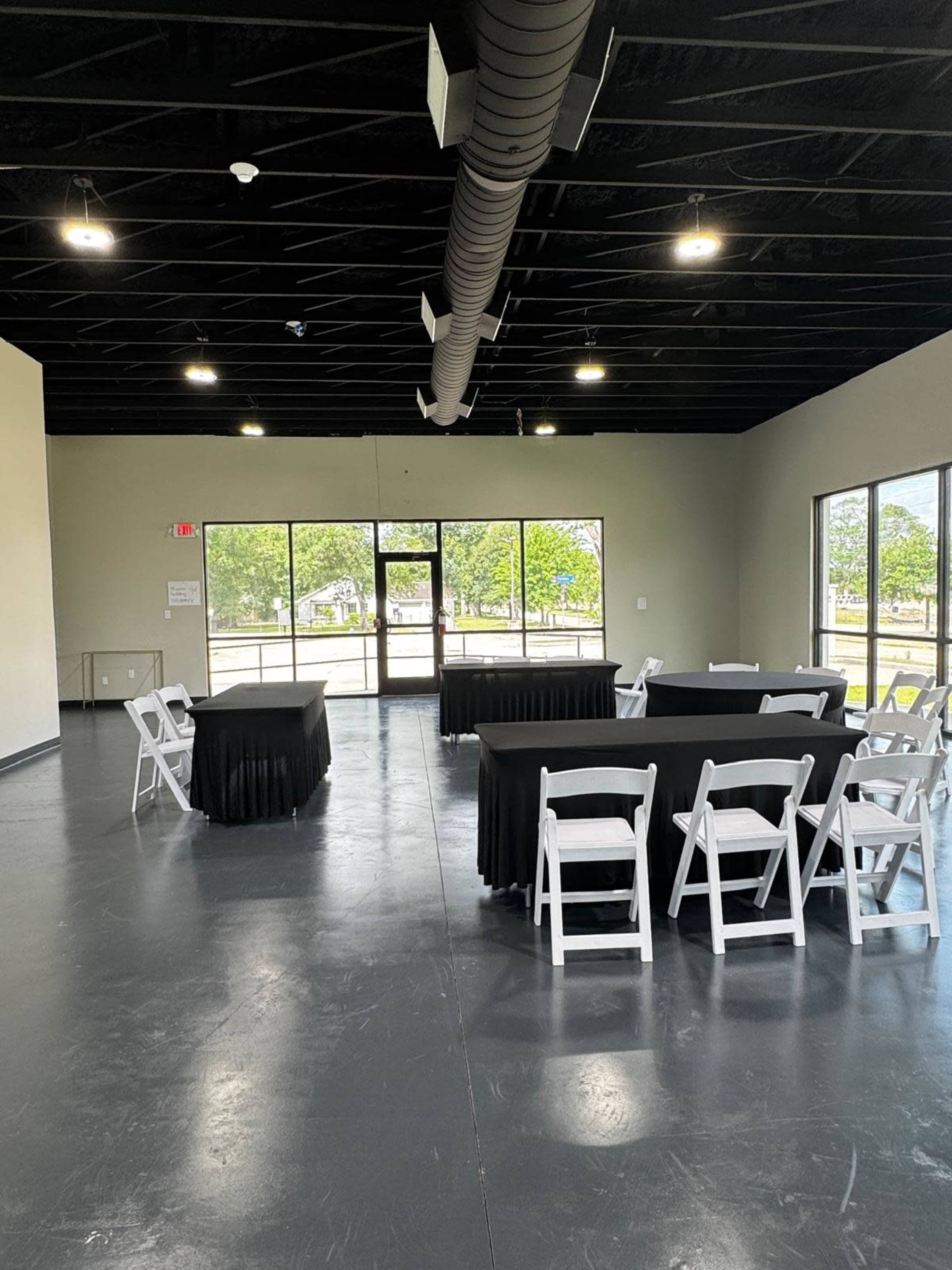The image shows a spacious, empty event room with several black tables and white chairs arranged neatly, and large windows letting in natural light.