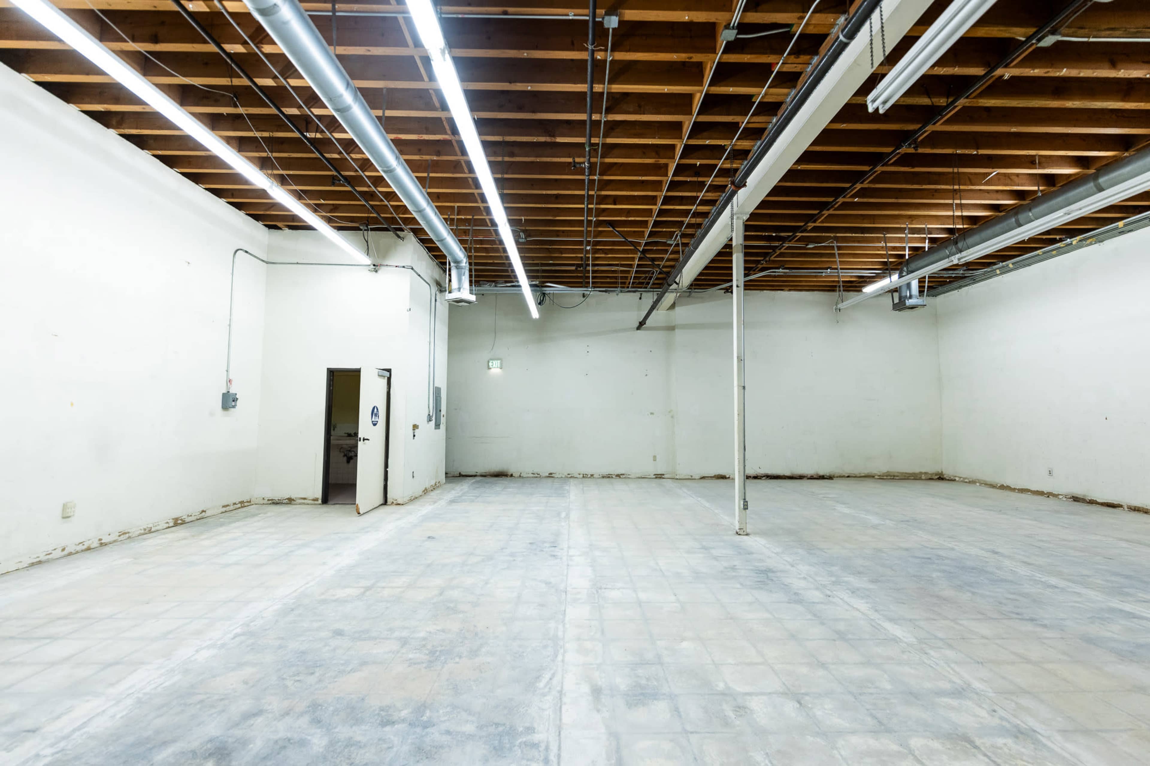The image shows a spacious, empty room with bare walls, a concrete floor, and exposed wooden beams overhead.