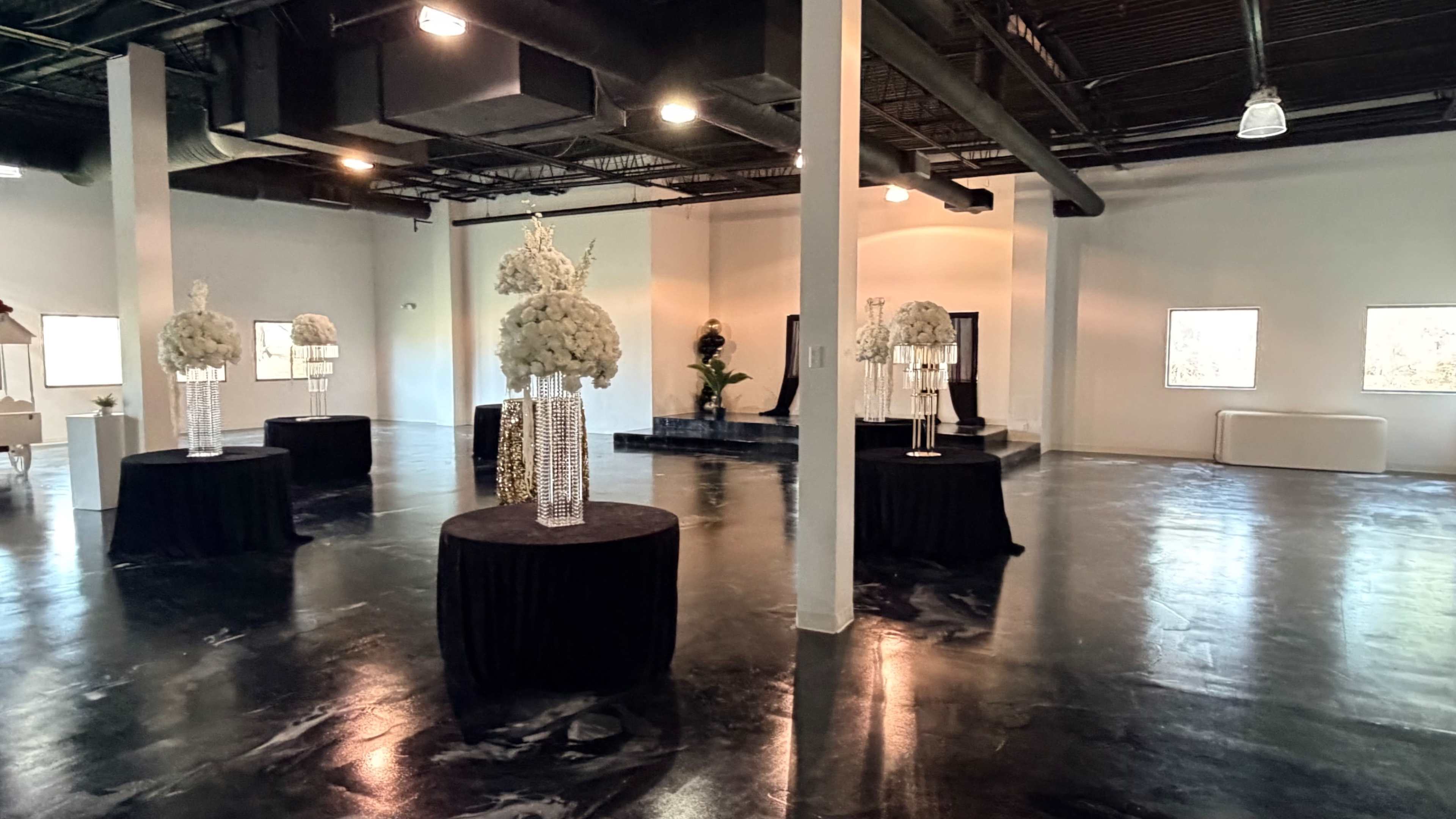 The image shows a spacious indoor venue with polished floors, featuring several black tablecloths with floral centerpieces arranged on stands.