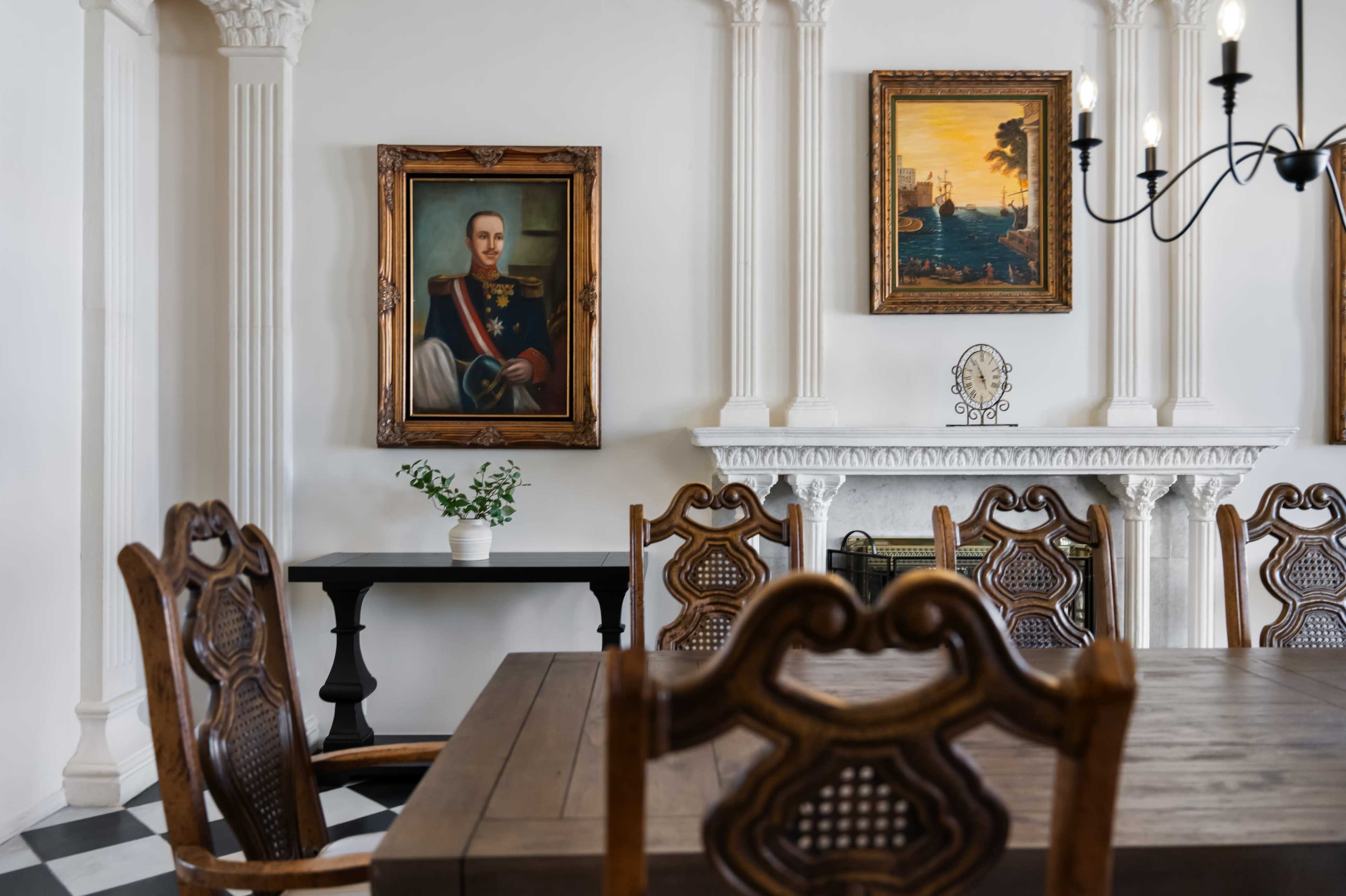 The image shows a wooden dining table surrounded by intricately designed chairs, with two framed paintings and a decorative plant displayed on the wall.