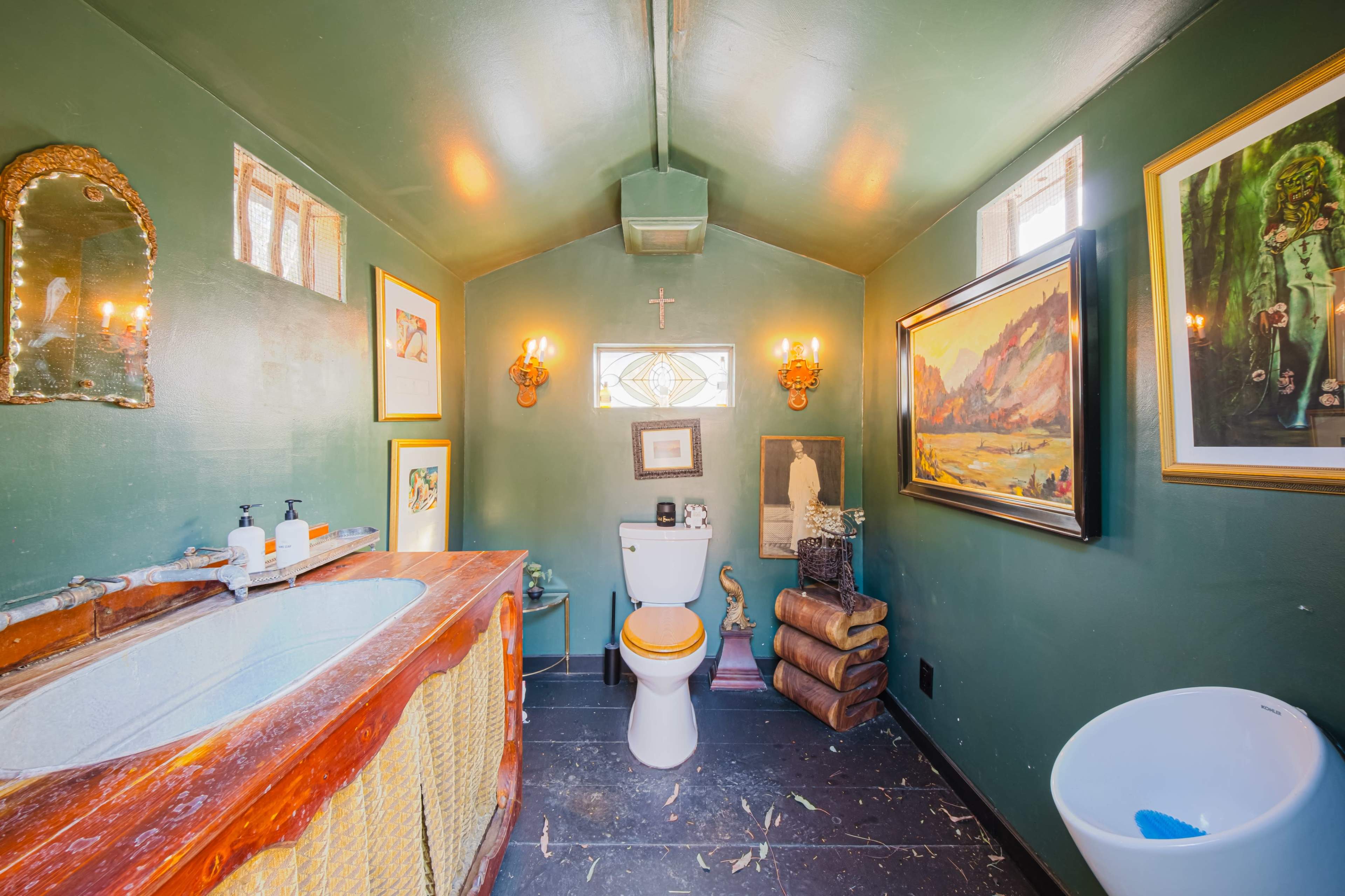 The image shows a small bathroom with dark green walls, featuring a wooden vanity, a white toilet, artwork on the walls, and windows providing natural light.