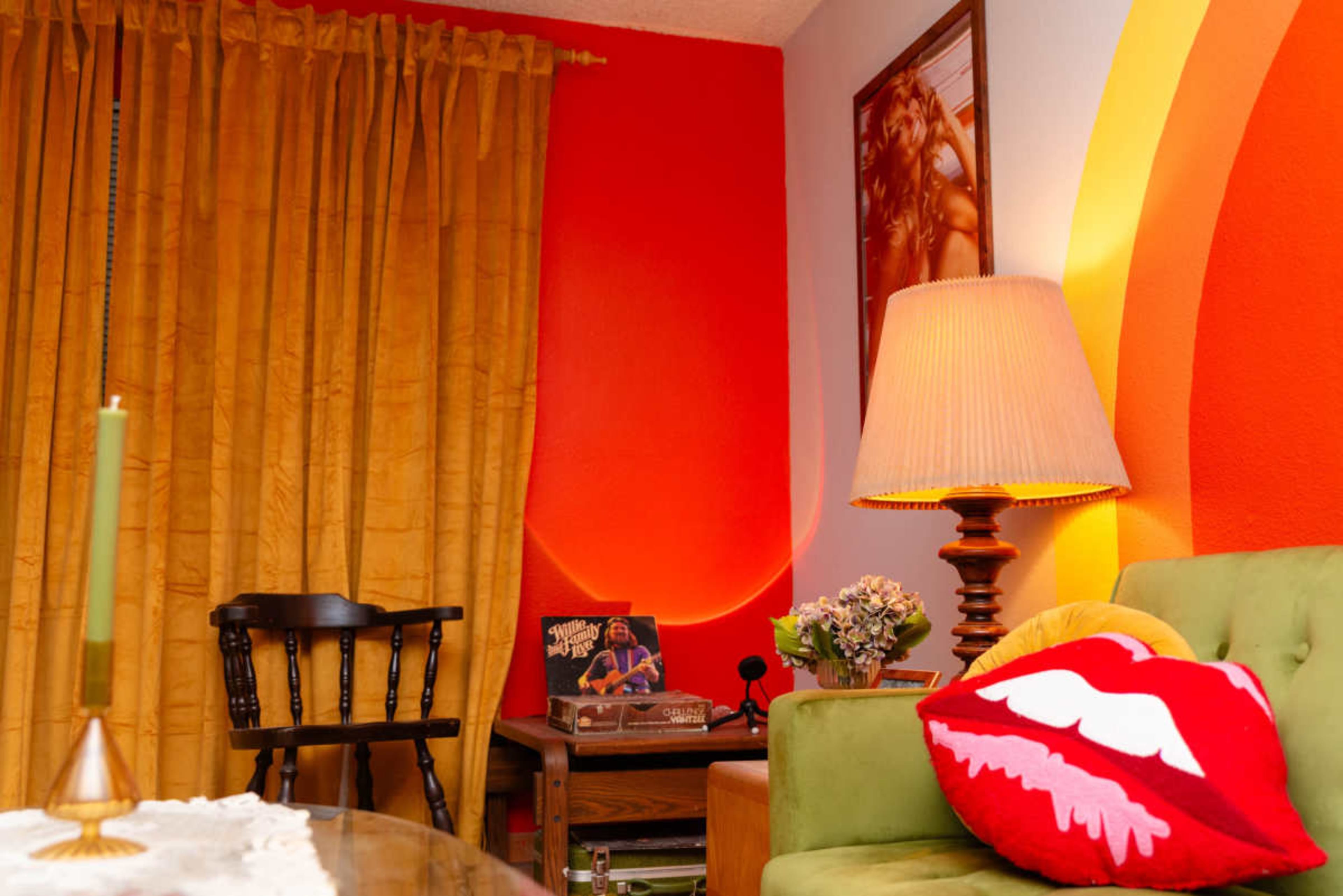 The image features a cozy living room with a green couch adorned with a lip-shaped pillow, a lamp on a side table, a wooden chair, and orange and yellow wall decorations.