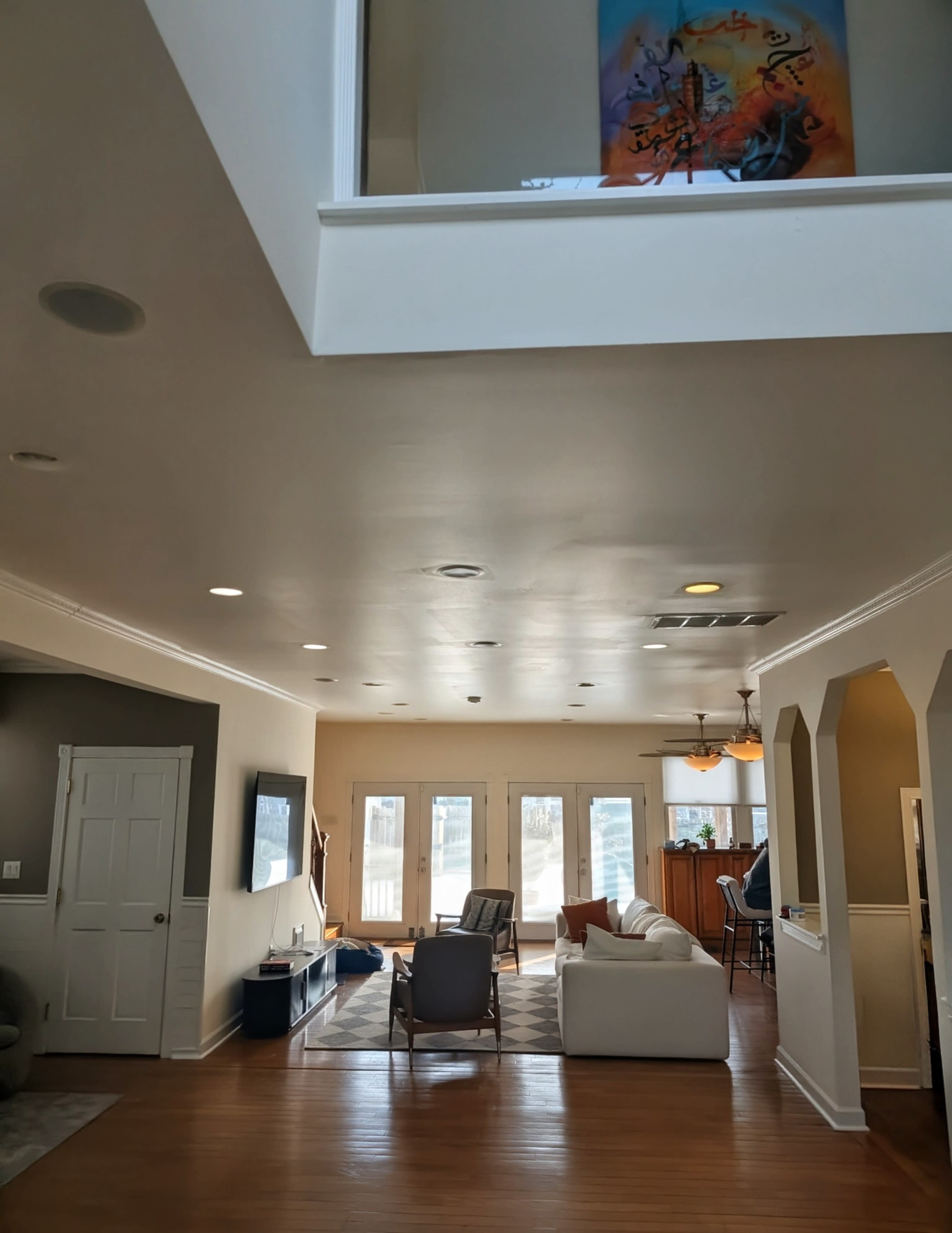 The image shows a spacious living area with a view of a kitchen, featuring large windows and modern furniture.